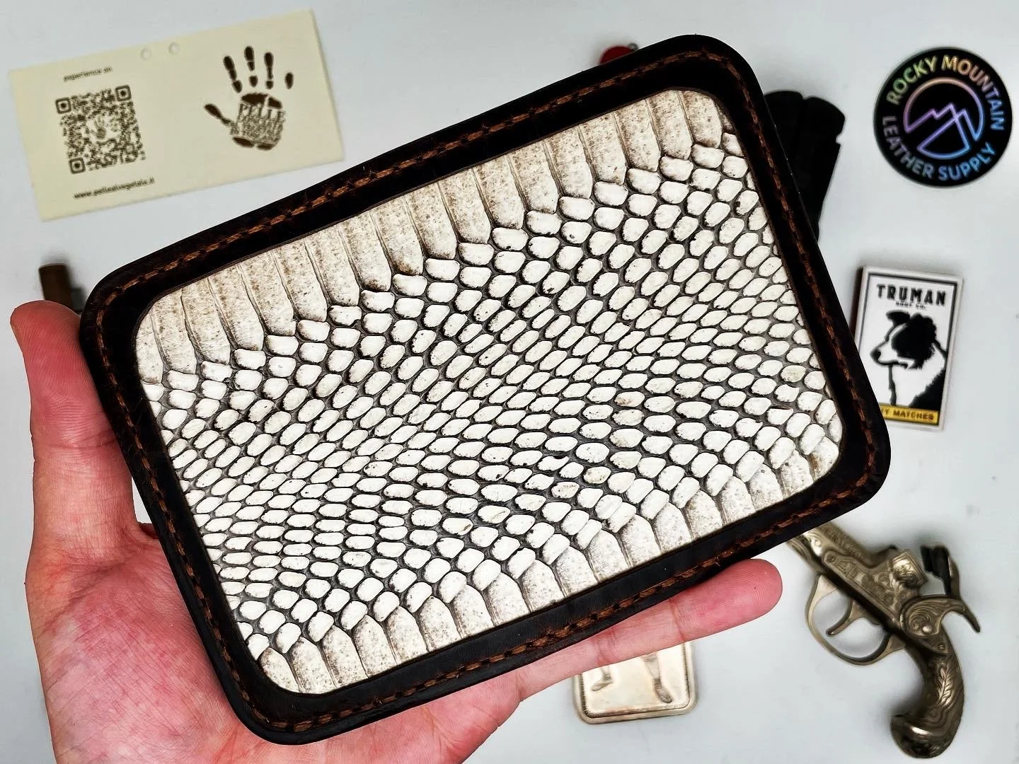 Hand holding a rectangular leather wallet with a snakeskin pattern on the surface, surrounded by a few stickers, a small gun, and other miscellaneous items on a white surface.