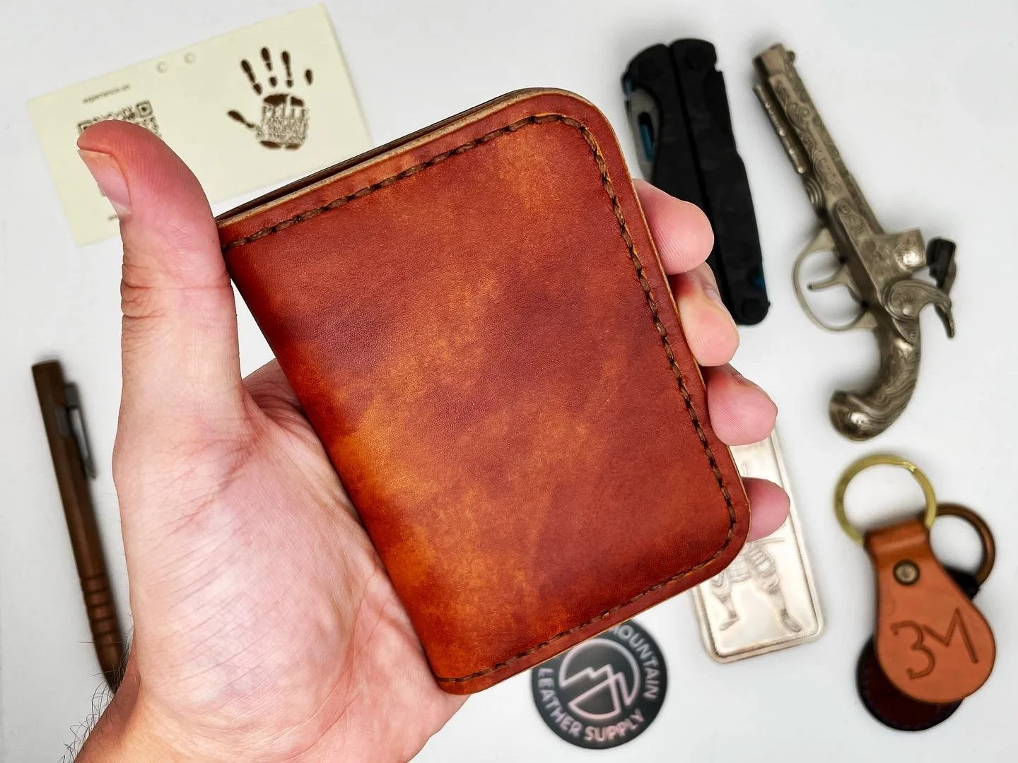 Hand holding a brown leather wallet with various items on a white surface in the background, including a toy gun, a lighter, a keychain, a piece of paper with a handprint, and stickers.