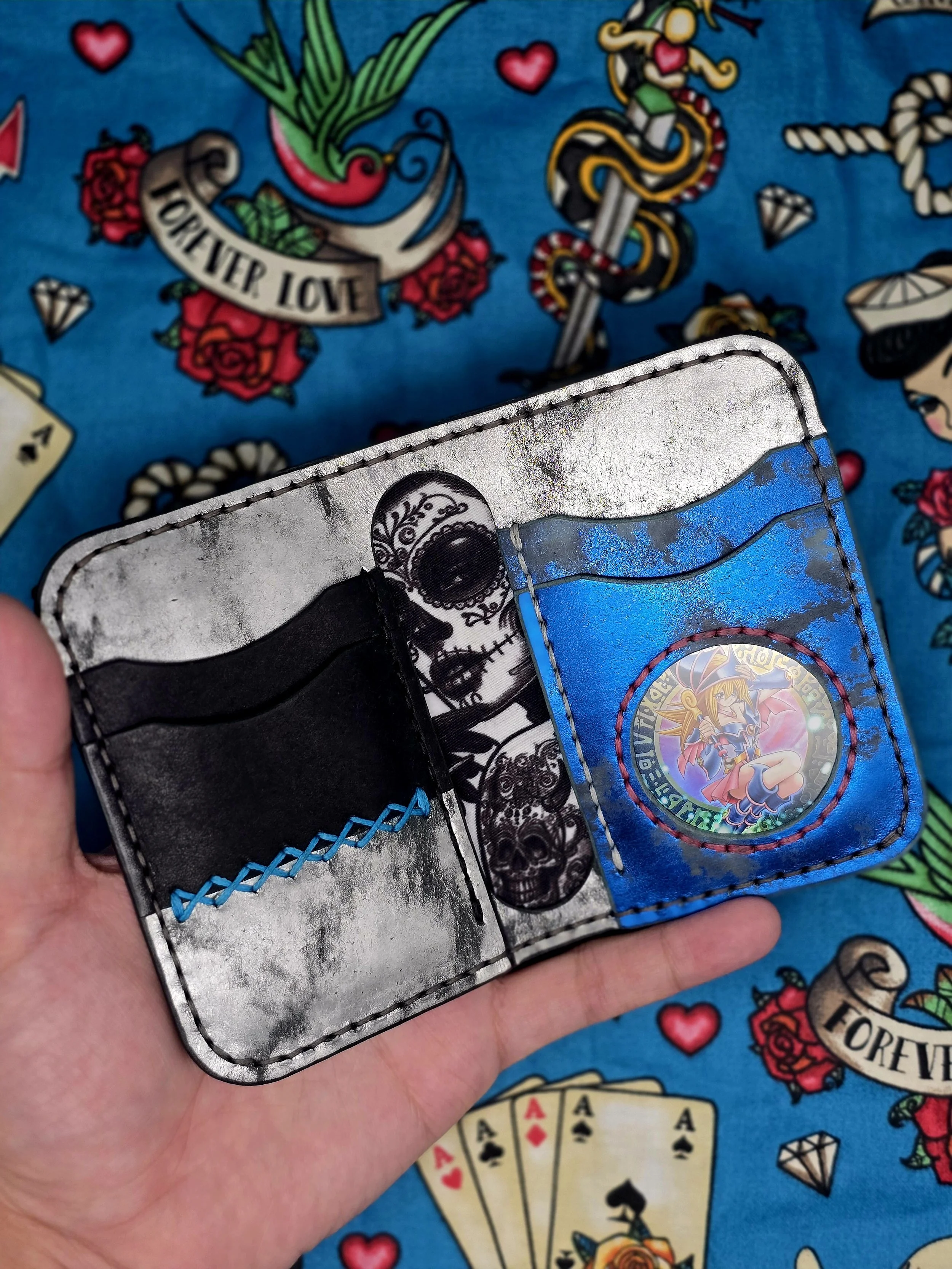 Open wallet with a black card, a TCG card featuring a character from Yu-Gi-Oh! and a skeleton design behind it, against a colorful background with tattoo-style illustrations and playing cards.