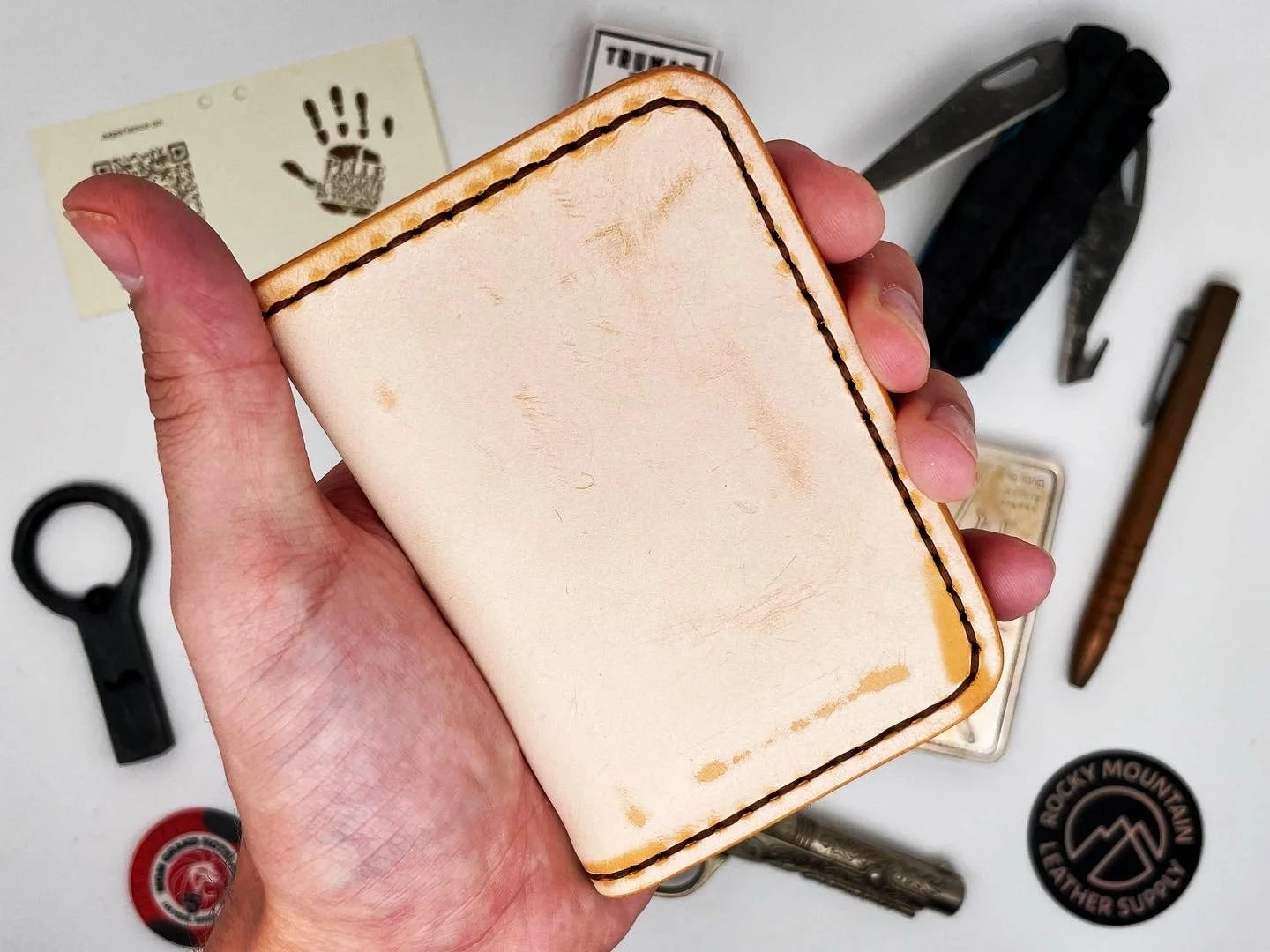 Hand holding a small, worn, blank beige leather wallet with dark stitching along the edges, surrounded by various tools and objects on a white surface, including a black multi-tool, a brown pocket knife, a small keychain, a circular item with a mount