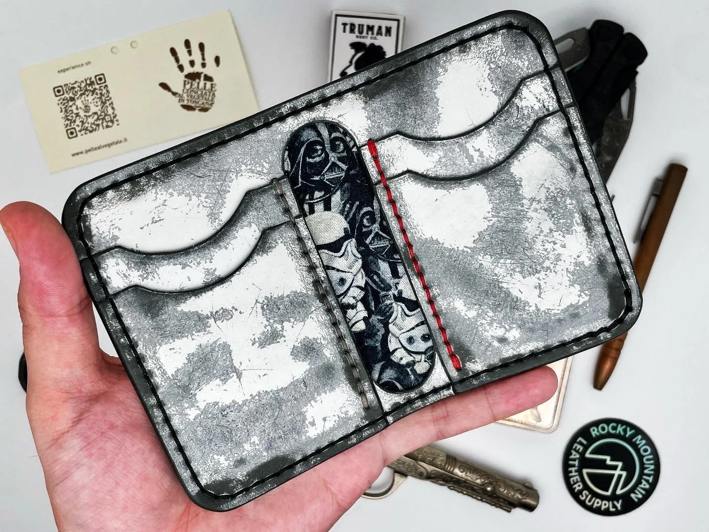 A metal wallet with a distress finish featuring an embroidered design of a person in black and white, with a red streak. The wallet is held by a person's hand over a white surface. Surrounding items include a Rocky Mountain Leather Supply sticker, a 