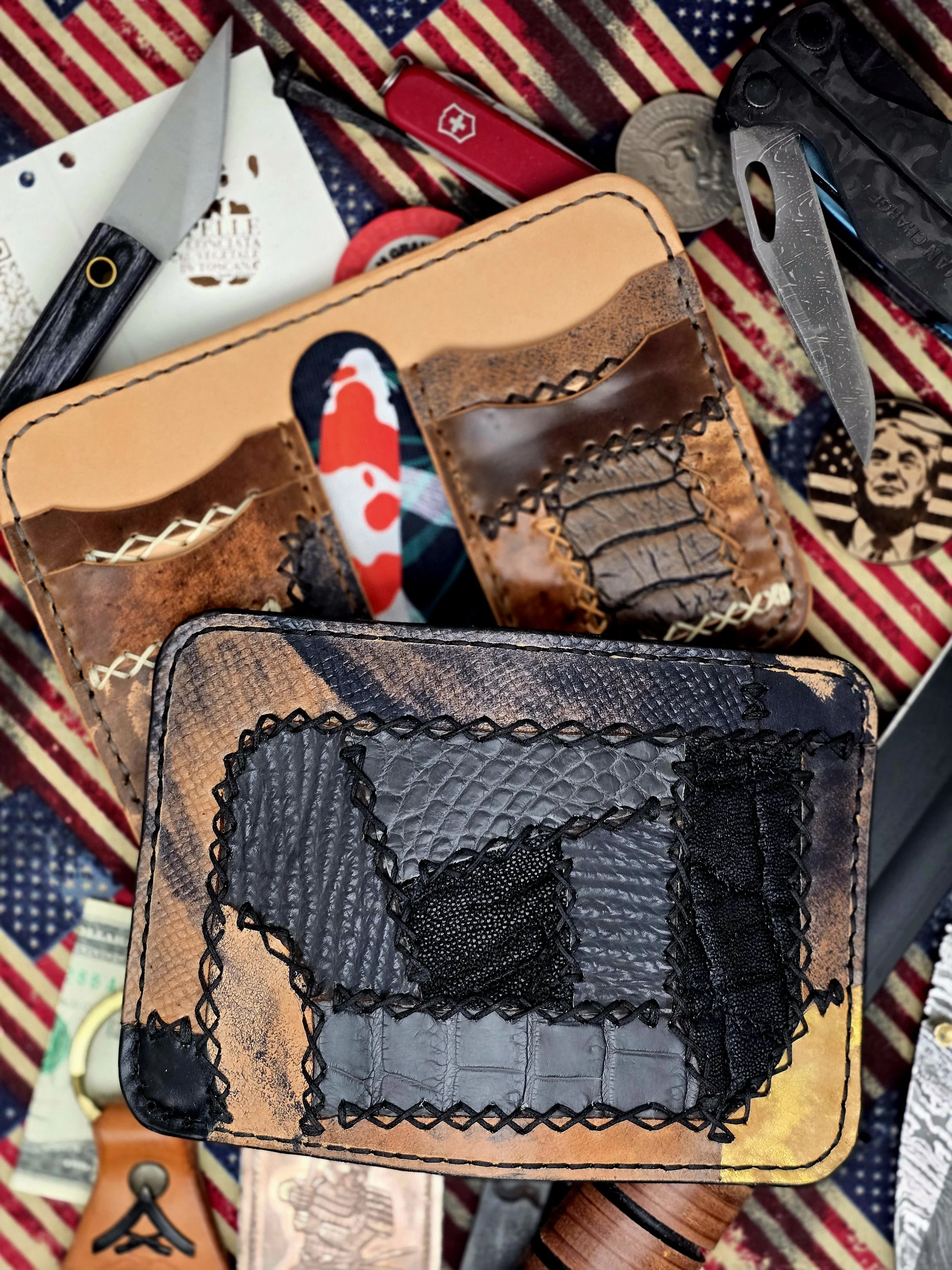Cowhide leather wallet with black fabric patch details, surrounded by knives, Swiss Army knife, coins, a tobacco tin, and dollar bills on a patriotic American flag fabric.