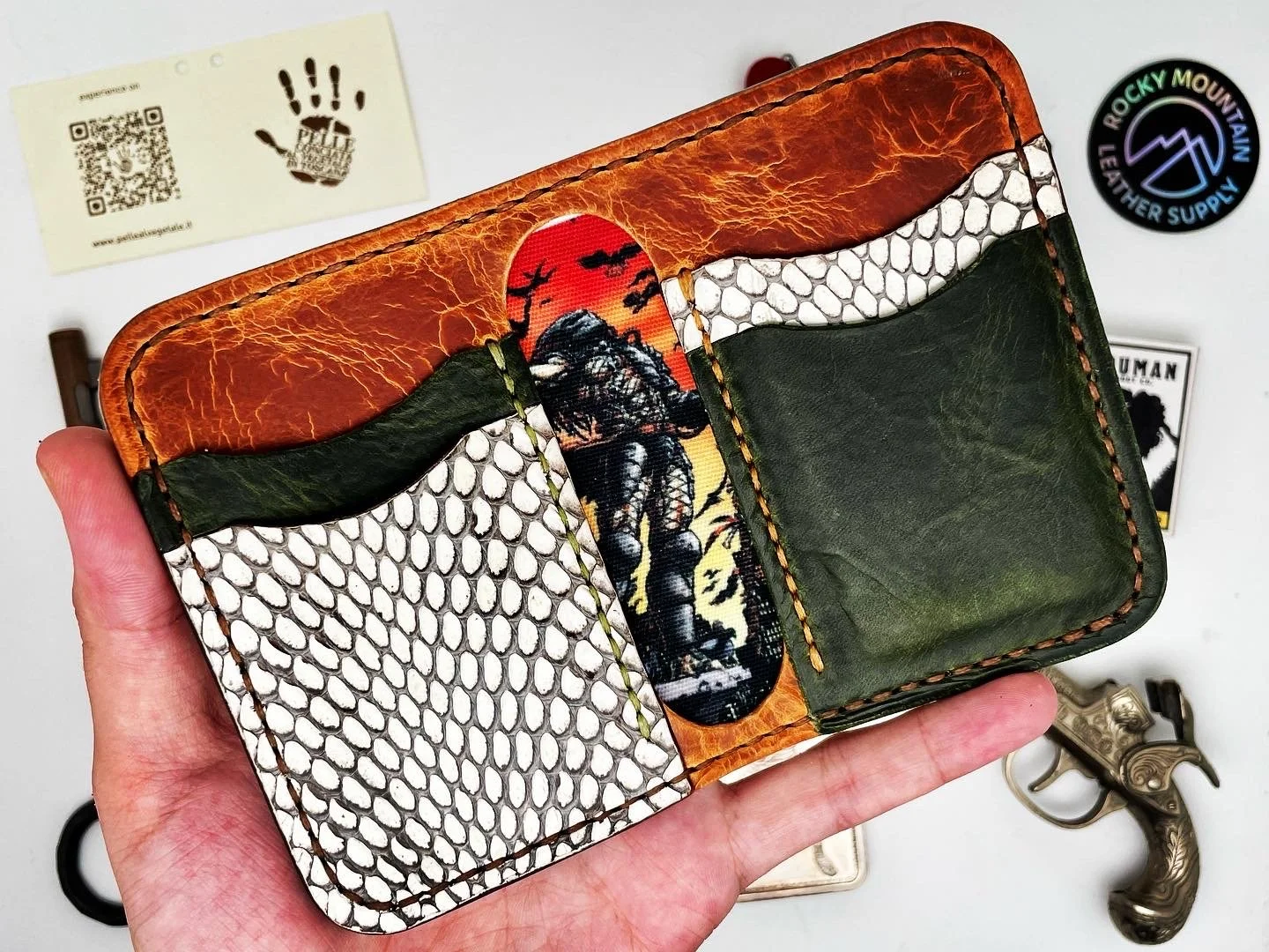 A leather wallet with a sunset and mountain scene, featuring snakeskin patterns and a green leather pocket, held in a person's hand. Surrounding items include a Rocky Mountain leather supply sticker, a tattoo business card, and a decorative pistol.