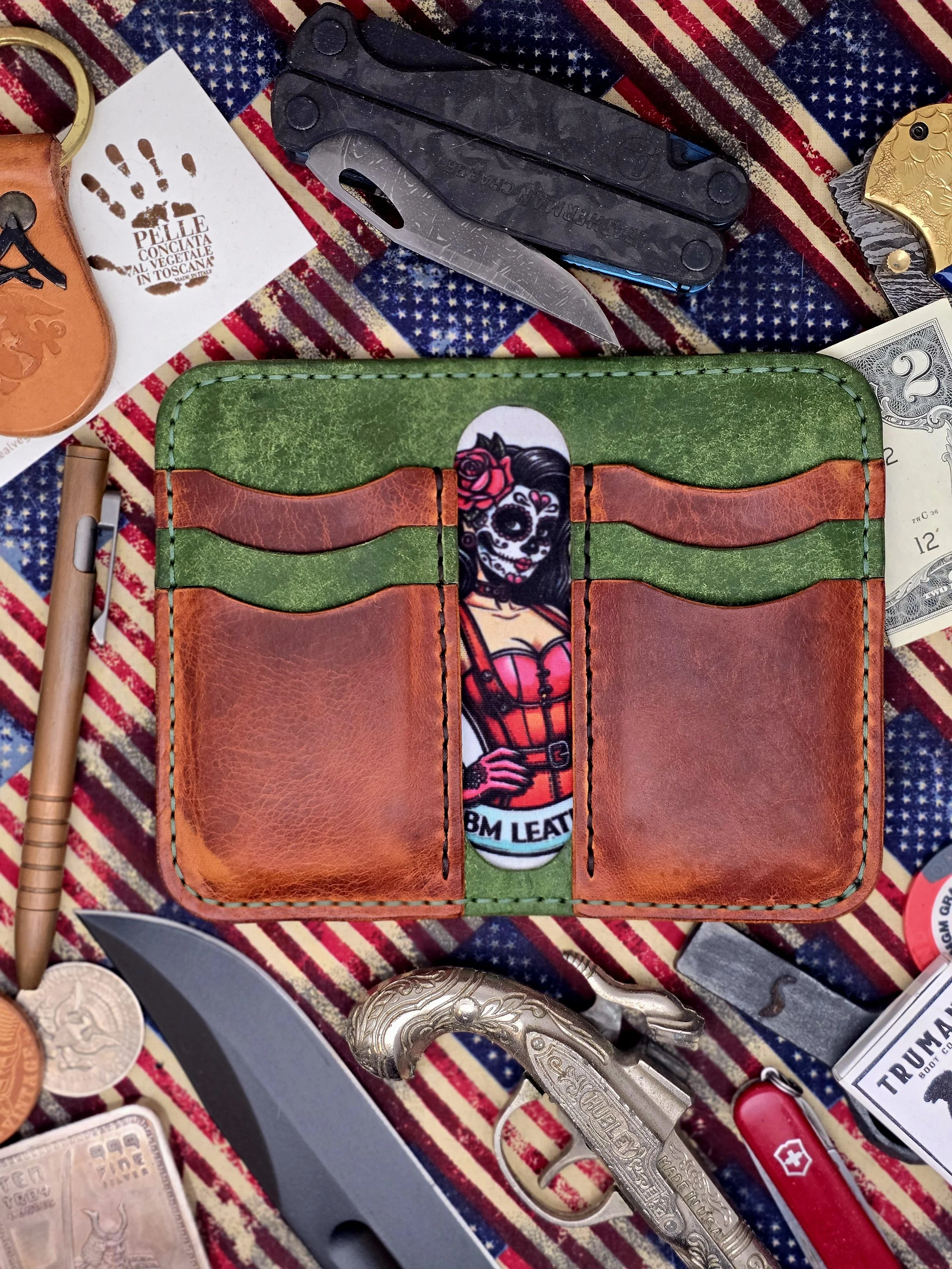 A leather wallet with a colorful illustration of a woman with a sugar skull face paint, wearing a red dress and a rose in her hair, set on a background of an American flag pattern alongside knives, coins, a lighter, and other miscellaneous items.