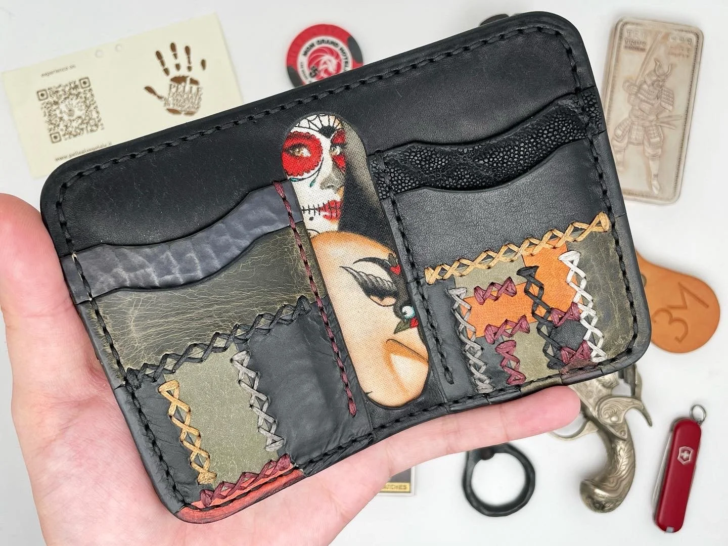 A black leather wallet with colorful patches and zigzag stitching, held in hand over a white surface with various objects including patches, a knife, a small pistol handle, and a paper with a QR code.