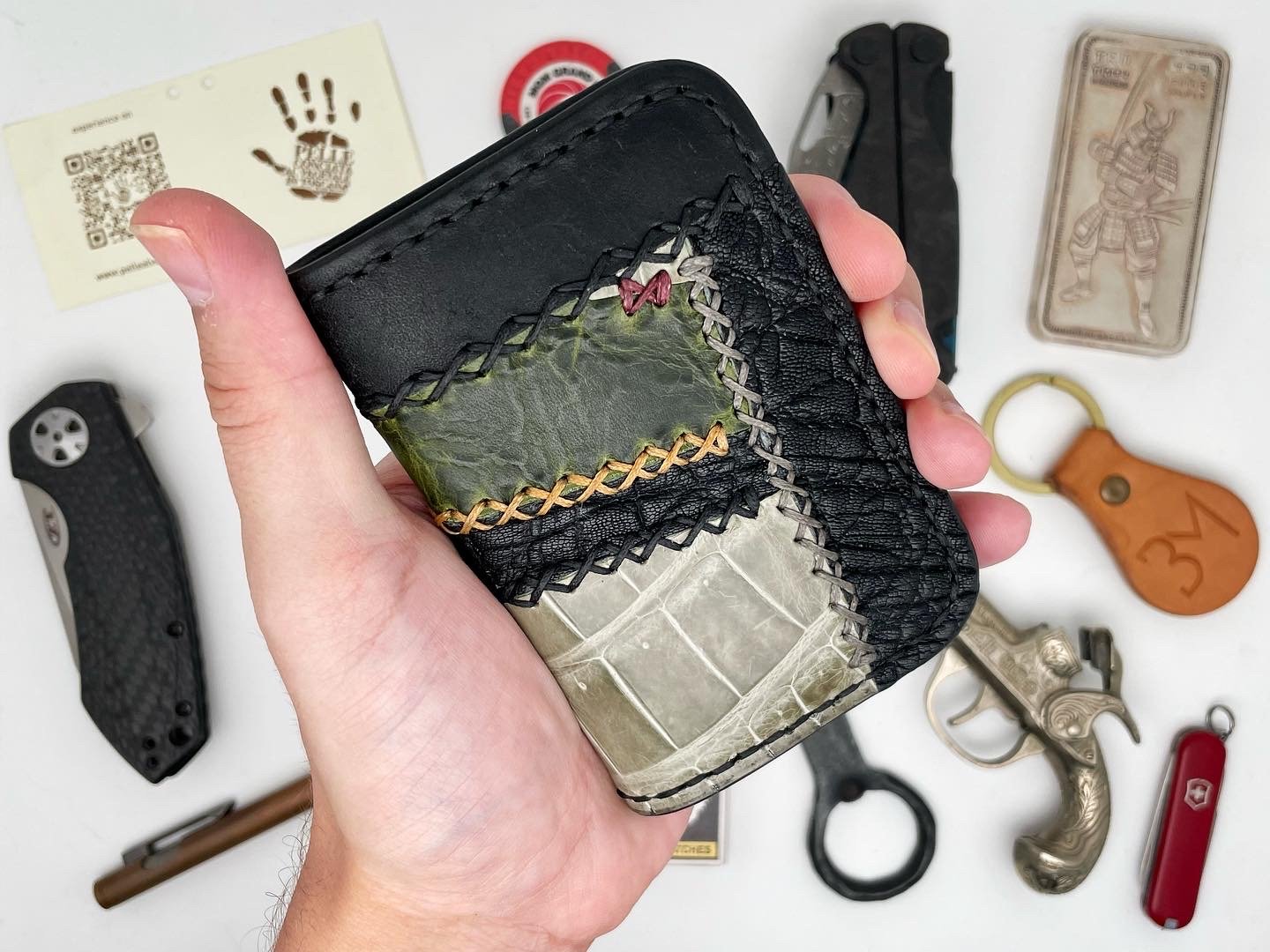 Close-up of a hand holding a black leather wallet with patches of colorful textured material on the front. Surrounding items on a white surface include a black pocket knife, a set of keys with a leather keychain, a red Swiss Army knife, a small metal
