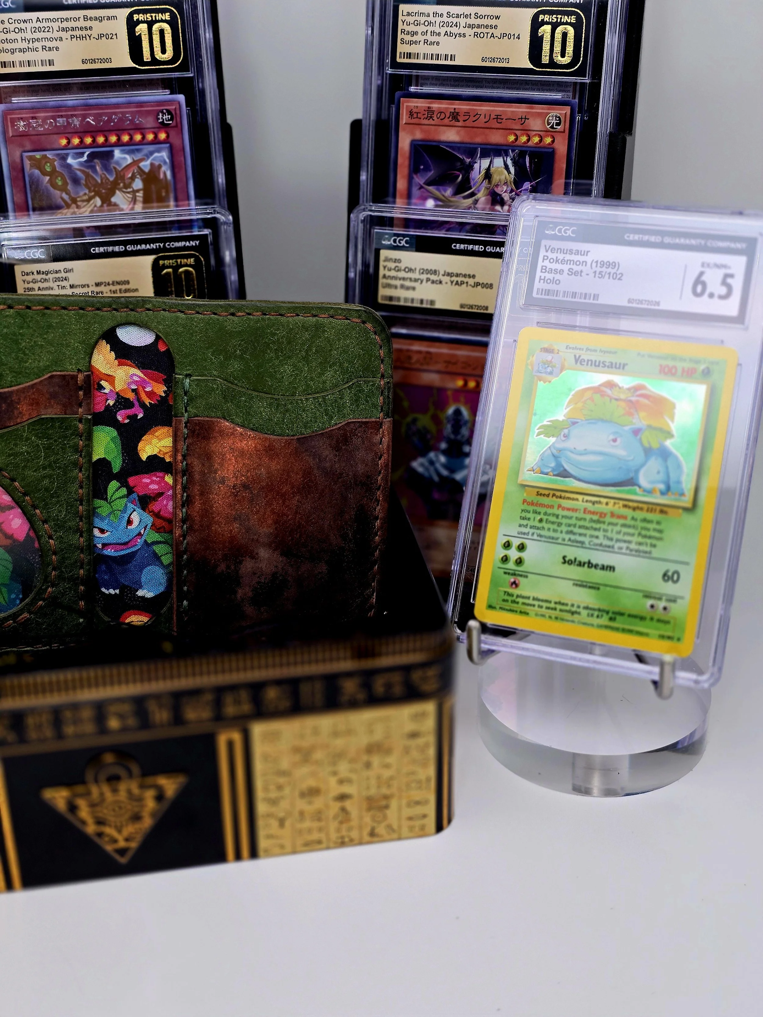Display of collectible cards including a Grade 6.5 graded Pokémon Venusaur card in a clear case and two Yu-Gi-Oh! cards graded 10 in protective cases, with a blurred Pokémon-themed wallet in the foreground.