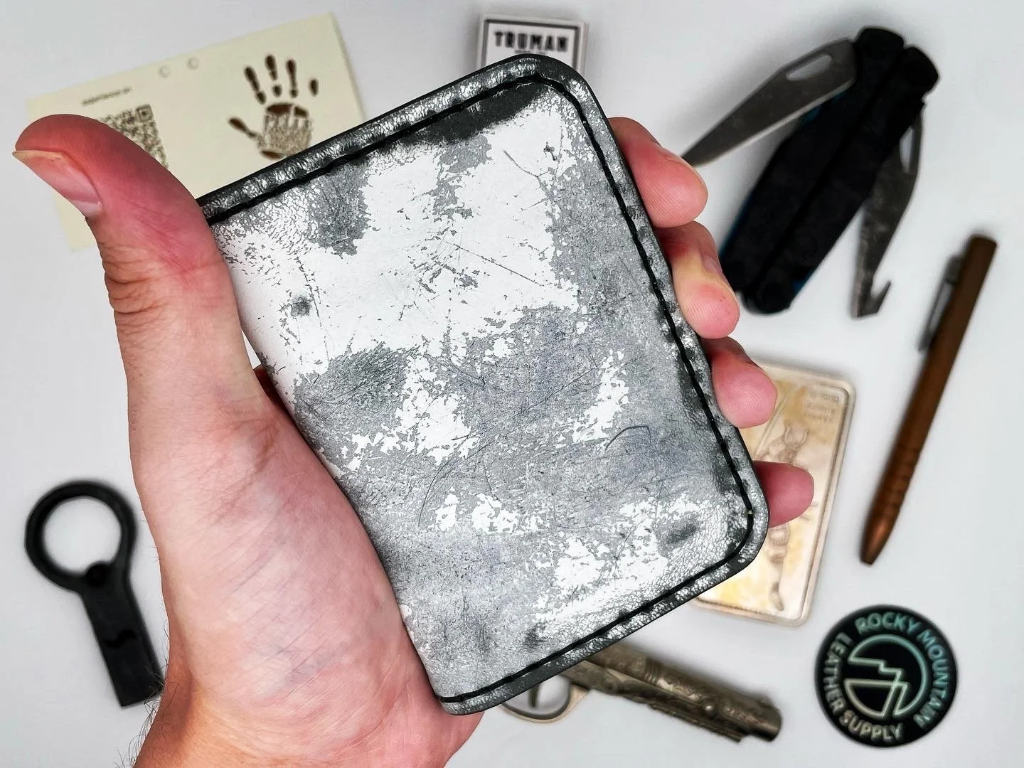 Hand holding a worn, metallic silver wallet with scratches and peeling paint, surrounded by various tools and items including a bottle opener, a baton, a patch, and some cards on a white surface.