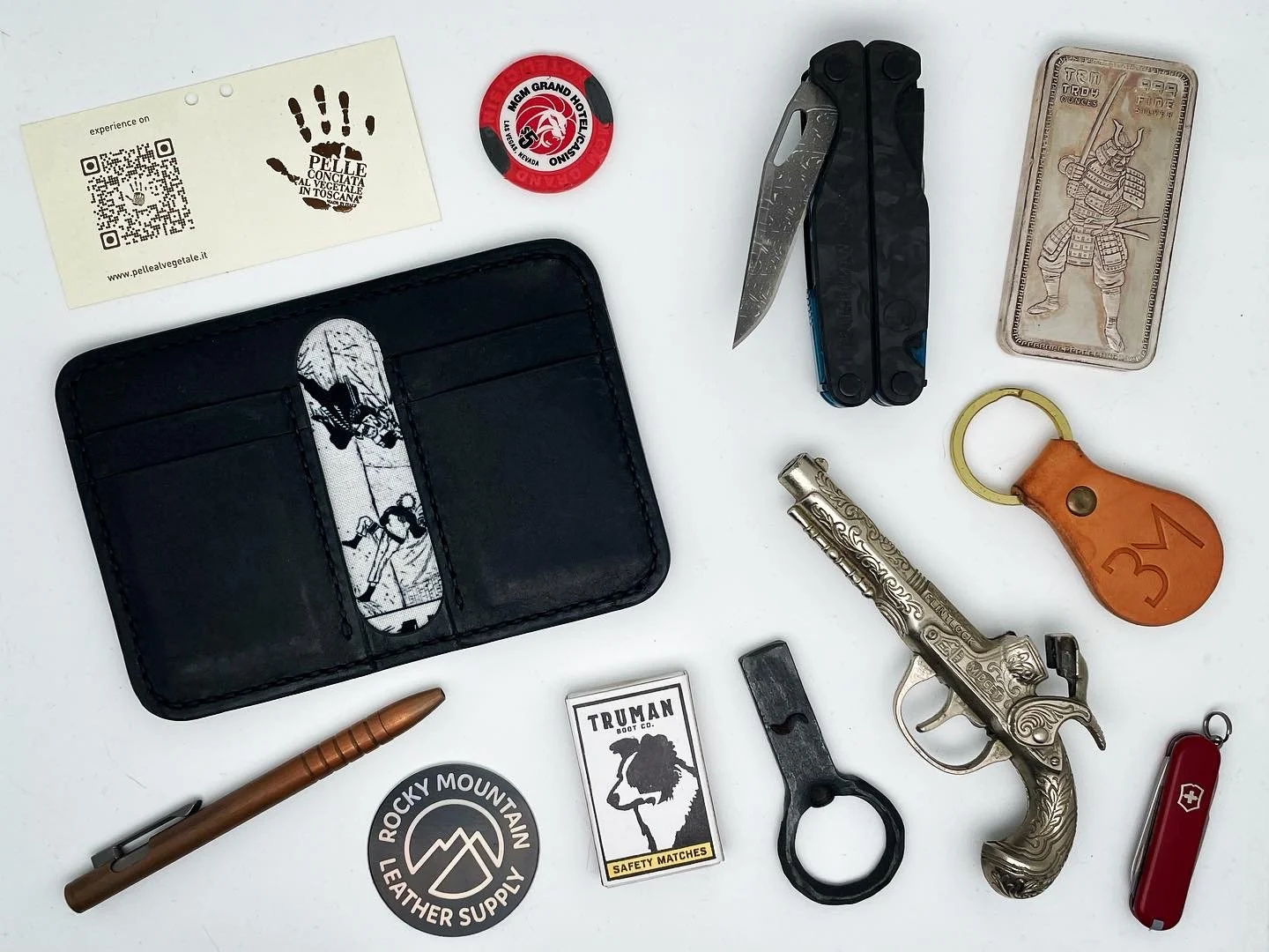 Collection of everyday items including a business card, a red and black round token, a black multitool, a silver firearm with ornate engravings, a red Swiss Army knife, a leather keychain, a small black tool, a packet of safety matches, a round stick