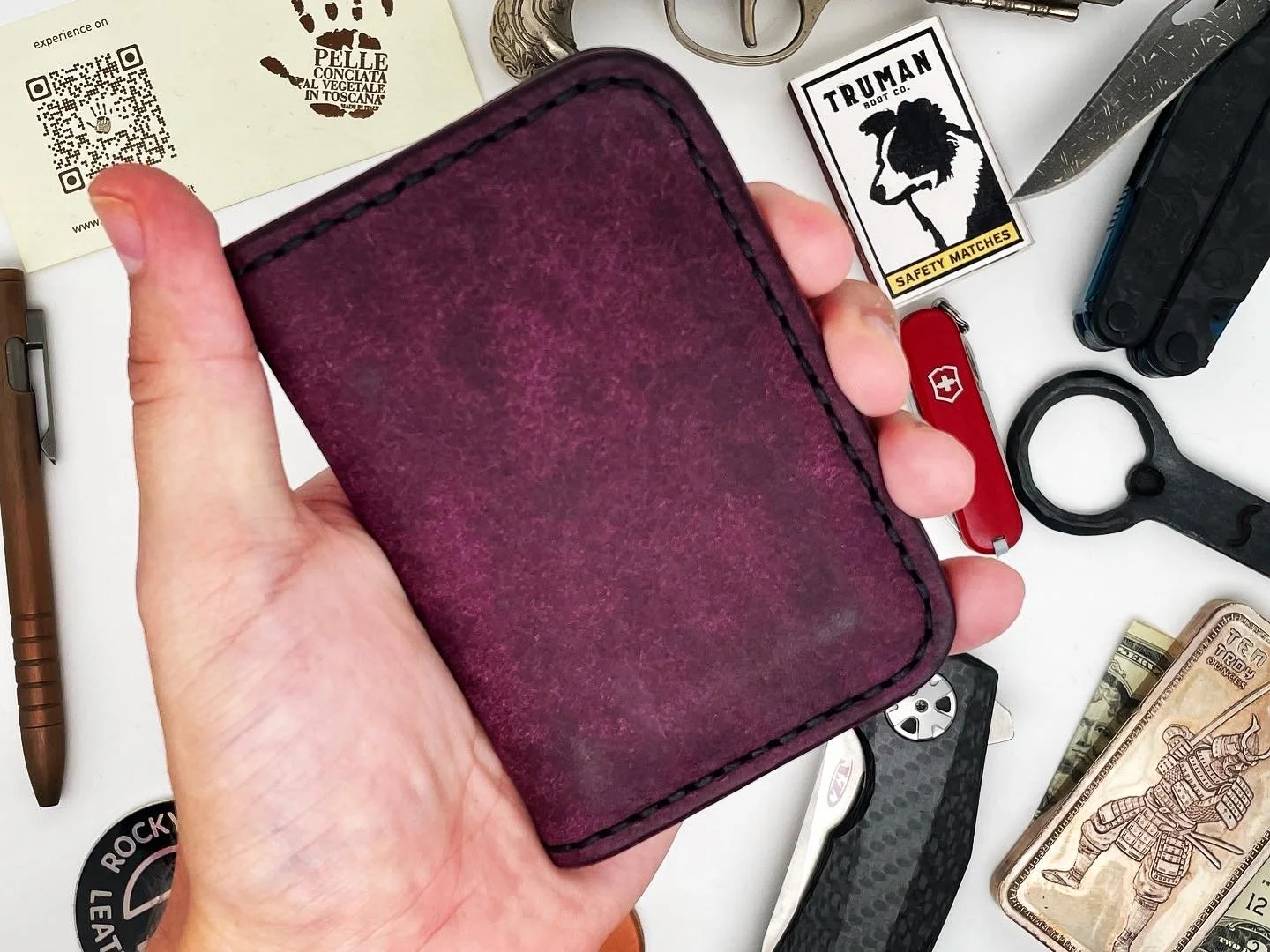 Hand holding a burgundy leather wallet surrounded by various items on a white surface, including a Swiss Army knife, pocket tools, a dollar bill, safety matches, a sticker, and a box.