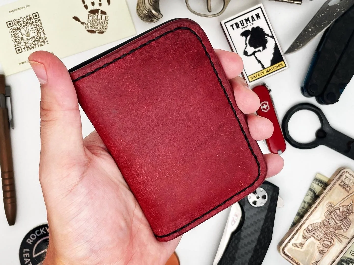 A hand holding a red leather wallet over a cluttered surface with various small items, including a Swiss Army knife, a black tactical tool, cash, a matchbox, and other miscellaneous objects.