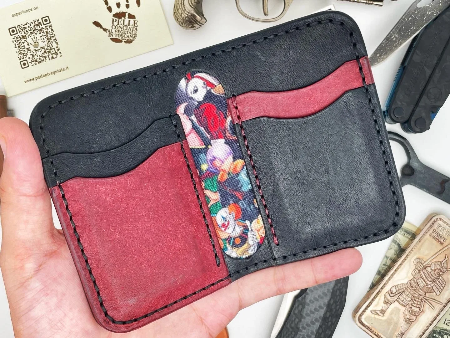 Hand holding a black and red wallet with a colorful cartoon character design on the inside tab. Various items including a Swiss Army knife, cash, and a card with a paw print logo are on the table.