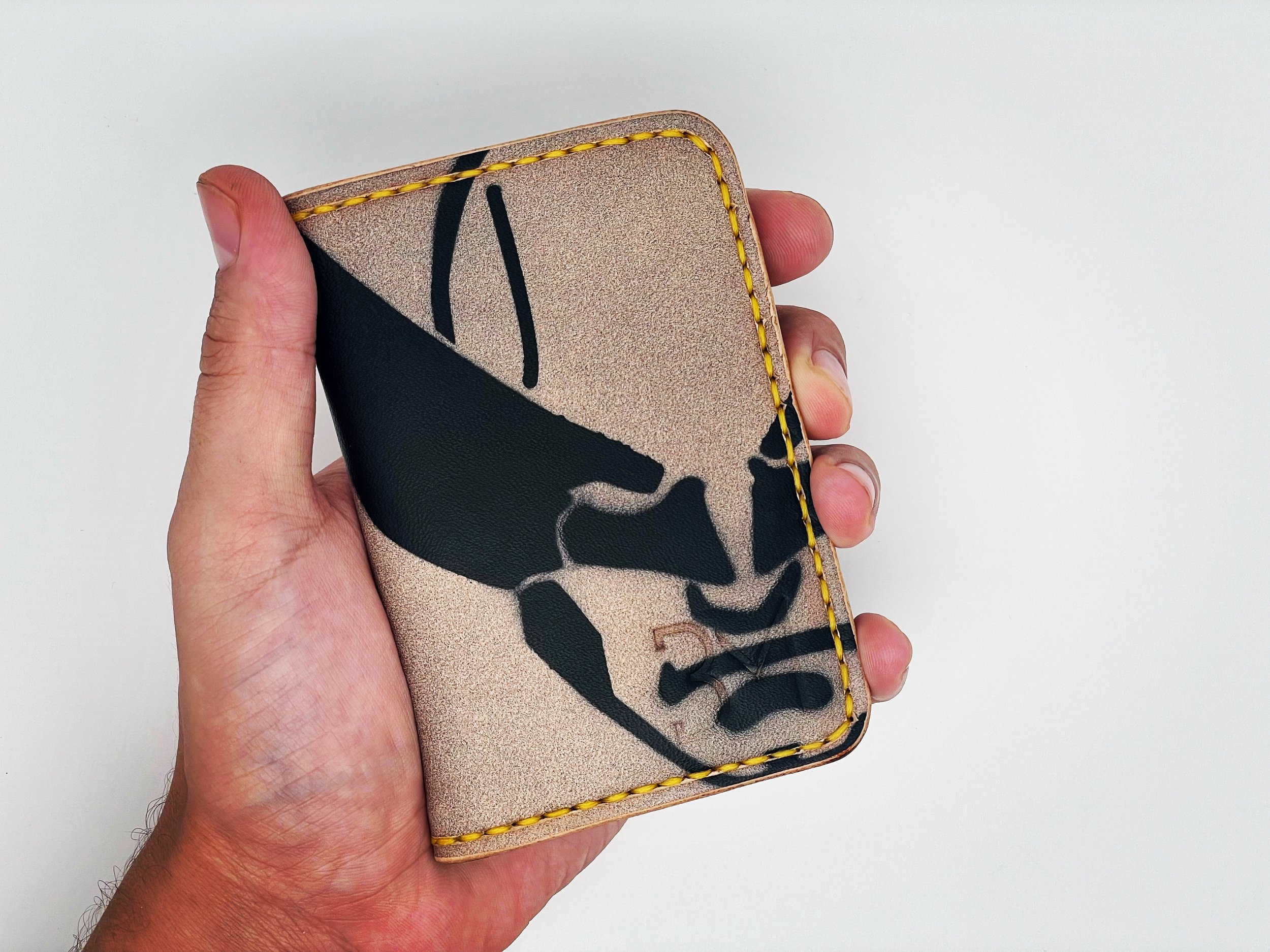 Hand holding a beige wallet with black, gold, and gray artistic design and yellow stitching on the edges, against a plain white background.