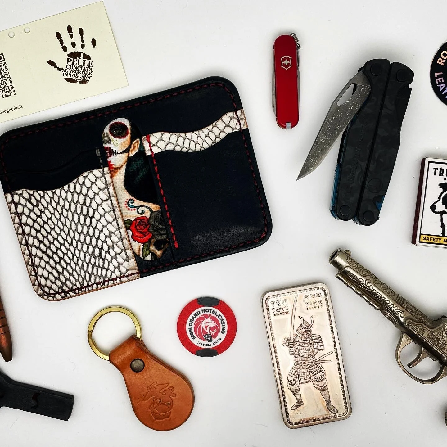 Collection of miscellaneous items on a white surface, including a comically decorated wallet, a red Swiss Army knife, a folding pocket knife, a stun gun, a metal lighter with an armored warrior design, a decorative leather keychain, a casino chip, an