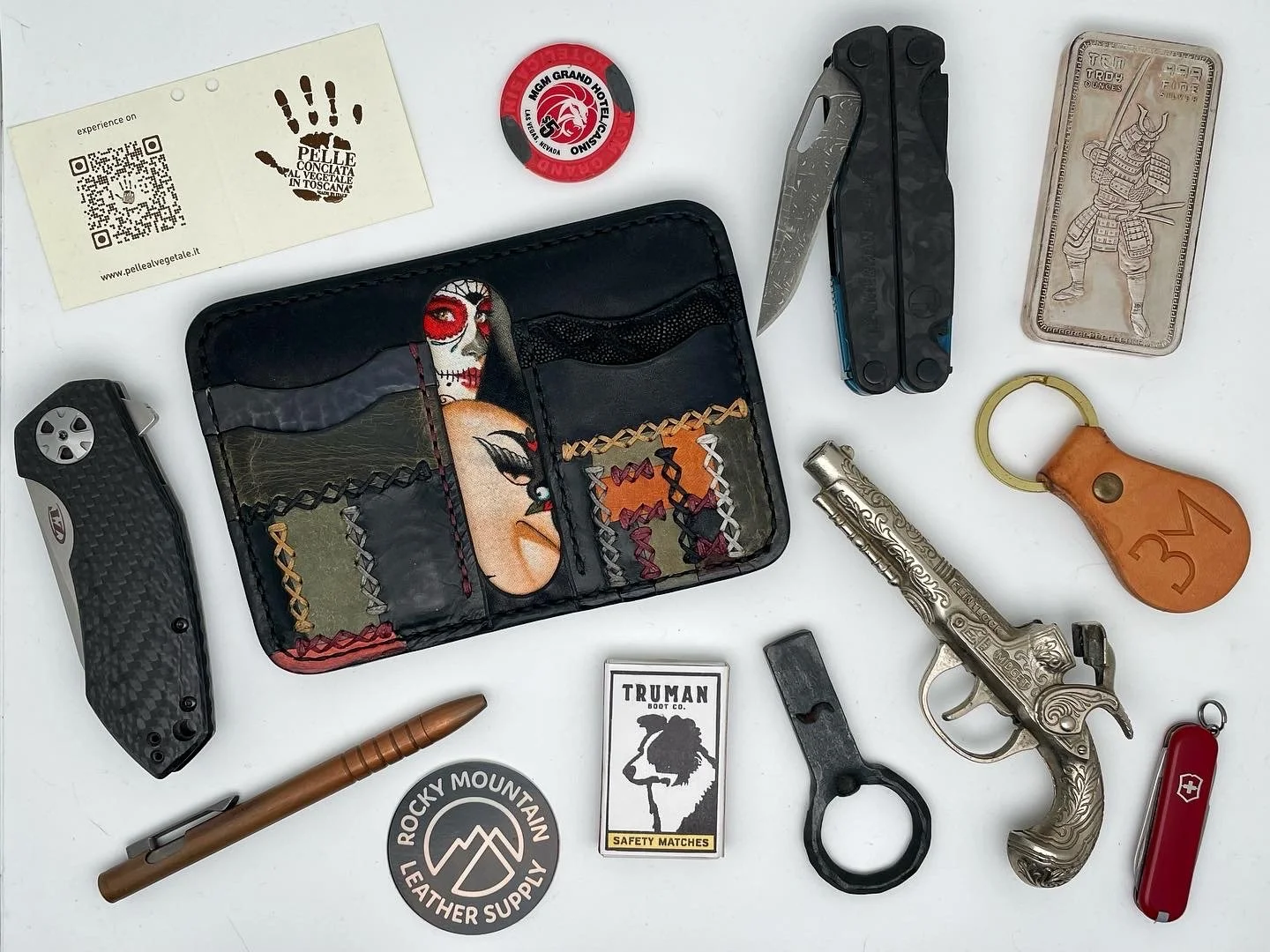 Assorted everyday carry items on a white surface, including a folding knife, a leather wallet with colorful design, a small pistol with engravings, a keychain with a leather cover, a red Swiss Army knife, a tube-shaped tool or flashlight, a matchbox 