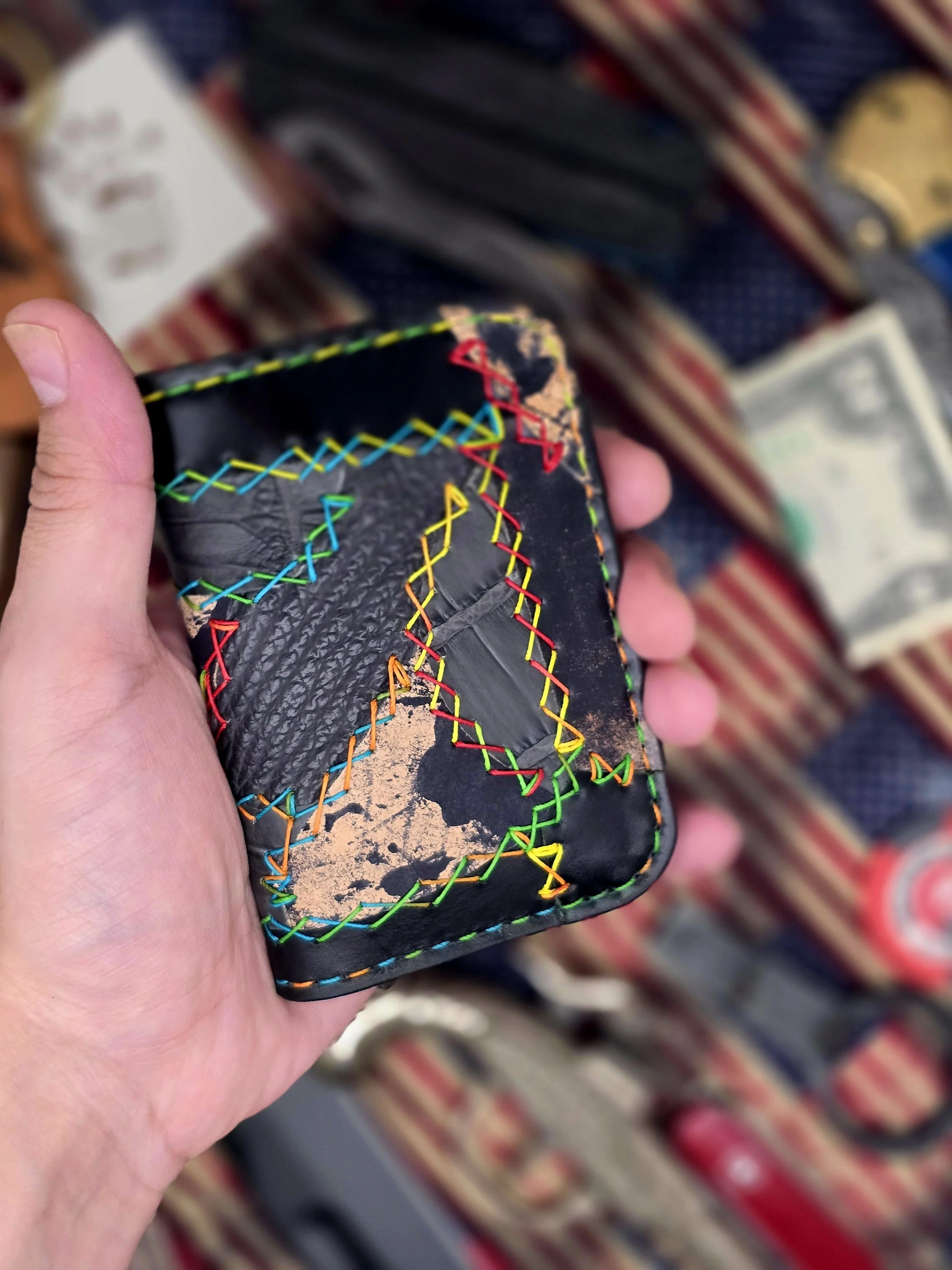 A black wallet with colorful stitched zigzag patterns and patches, held in a person's hand, with a background of striped fabric and some cash.