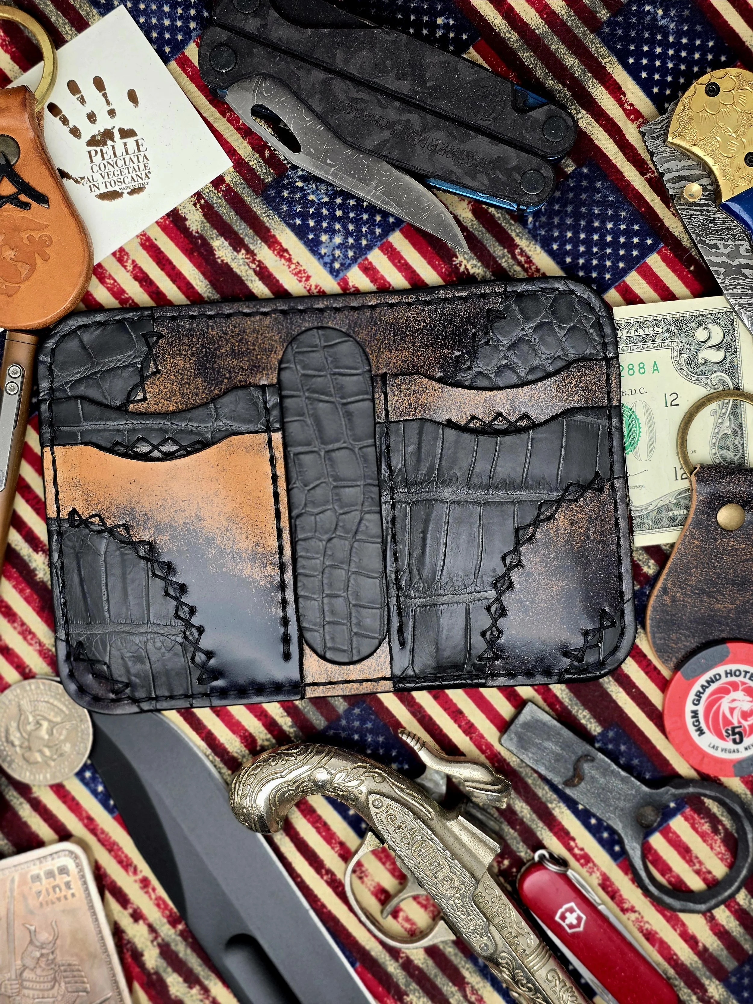 A collection of various knives, a leather wallet with a crocodile pattern, U.S. currency, and miscellaneous items on a table with an American flag pattern.
