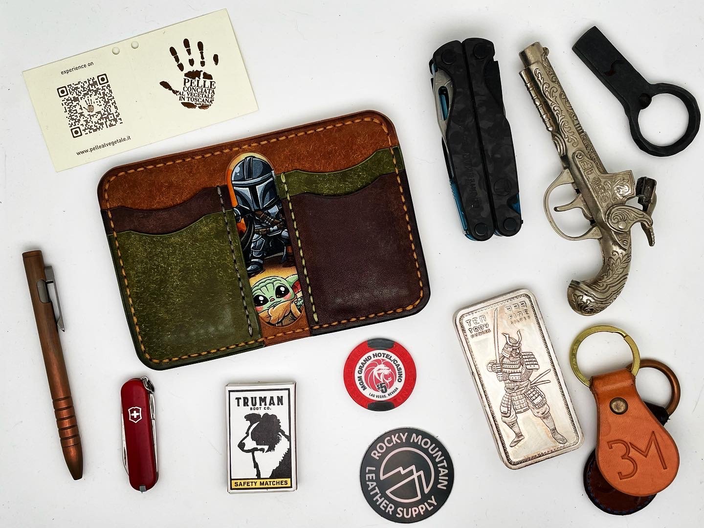Collection of everyday carry items including a leather wallet, a multifunction tool, a small pistol replica, a keychain with a leather tag, a red Swiss Army knife, a pen, a tile tracker, a metal coin, a silver bar, a safety matchbox, and a round toke