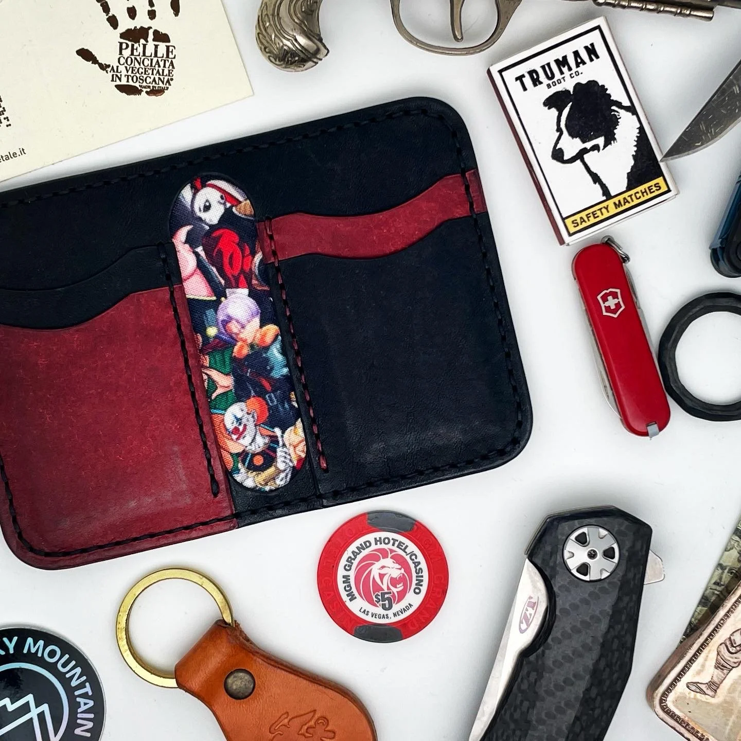 A collection of small everyday carry items on a white surface, including a black leather wallet with a colorful cartoon character on it, a Swiss Army knife, a pair of safety matches, a poker chip from MGM Grand Hotel Las Vegas, a small leather keycha
