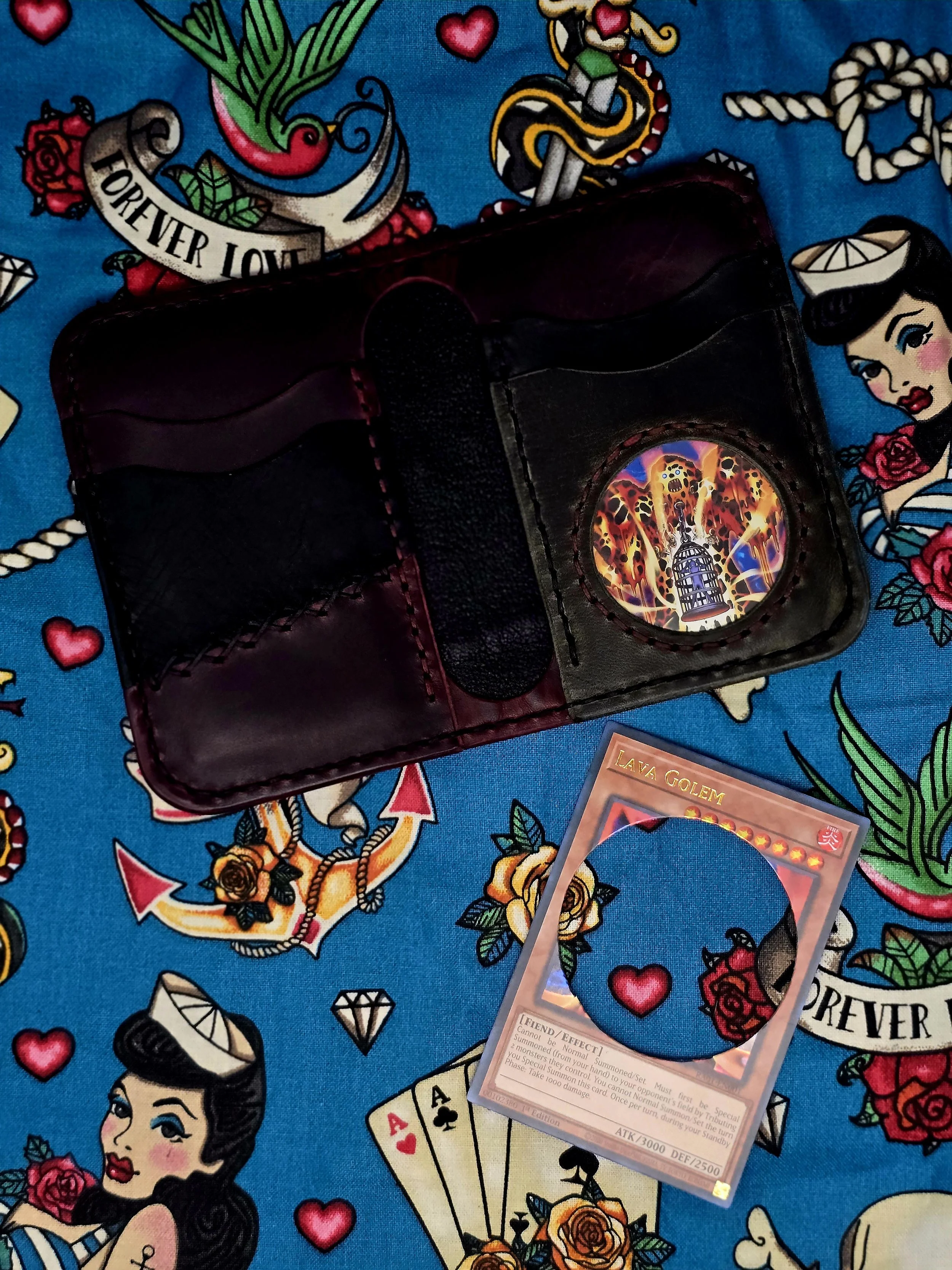 A leather wallet and a Yu-Gi-Oh! trading card, with the card titled 'Lava Golem,' resting on a colorful cloth with tattoo-style artwork featuring women, hearts, roses, and nautical themes.
