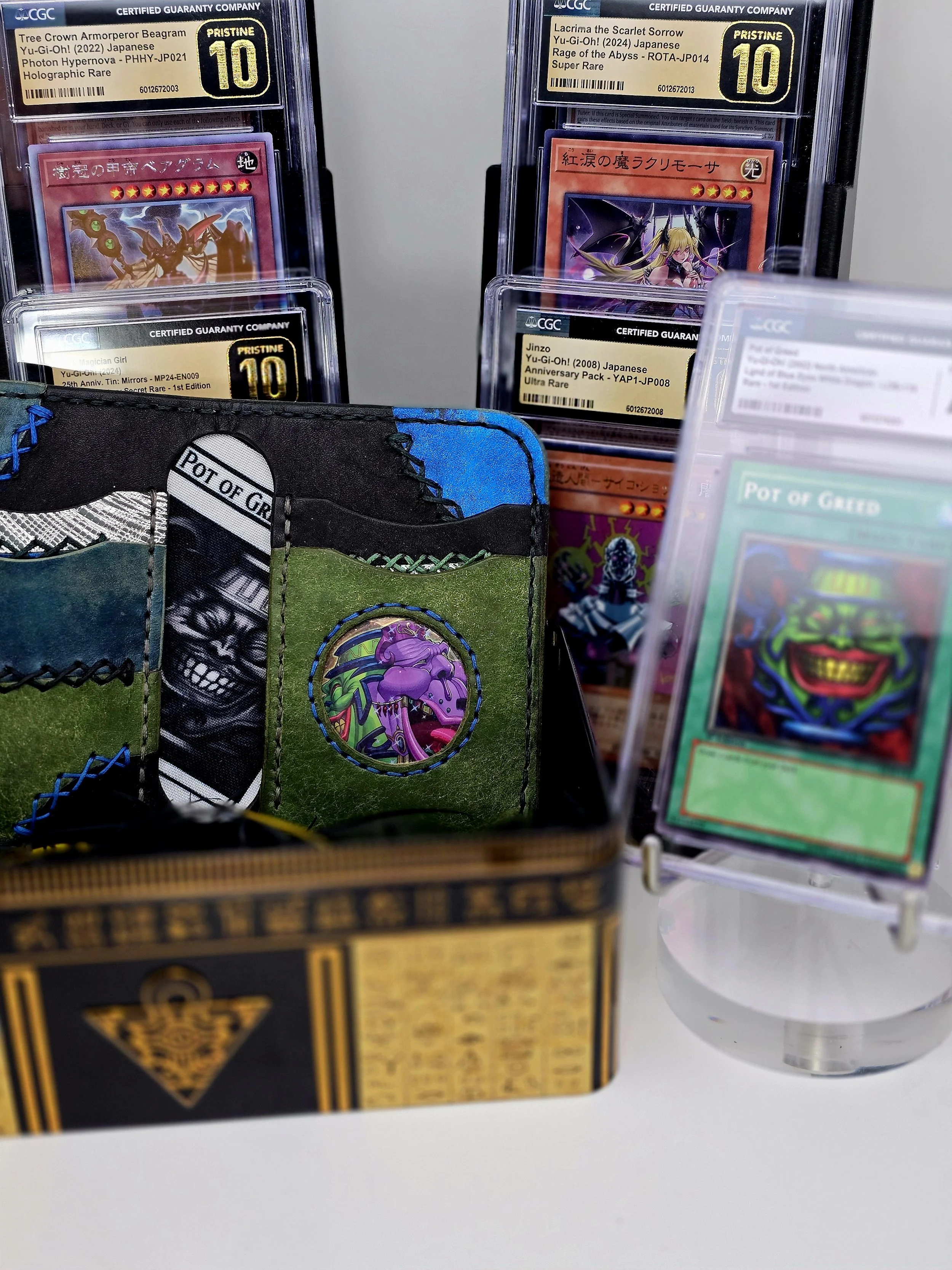 Close-up of Yu-Gi-Oh! trading cards and a deck box with artwork, including some cards in protective sleeves and a Pot of Greed card displayed on a stand.