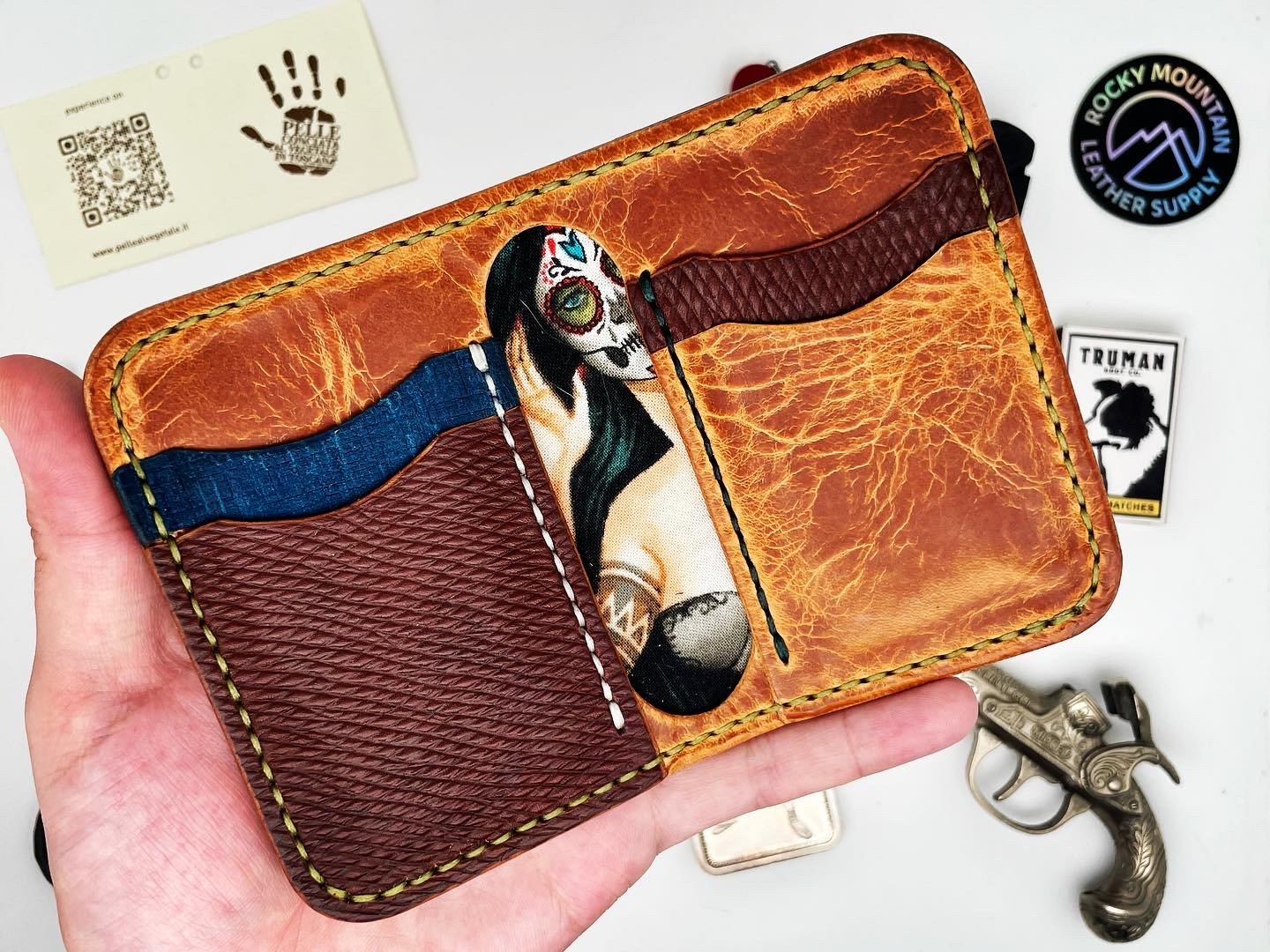 Hand holding a leather wallet with a painted woman with sugar skull face makeup, wearing a tank top, and long hair. Surrounding items include patches, stickers, and a small silver gun-shaped object.