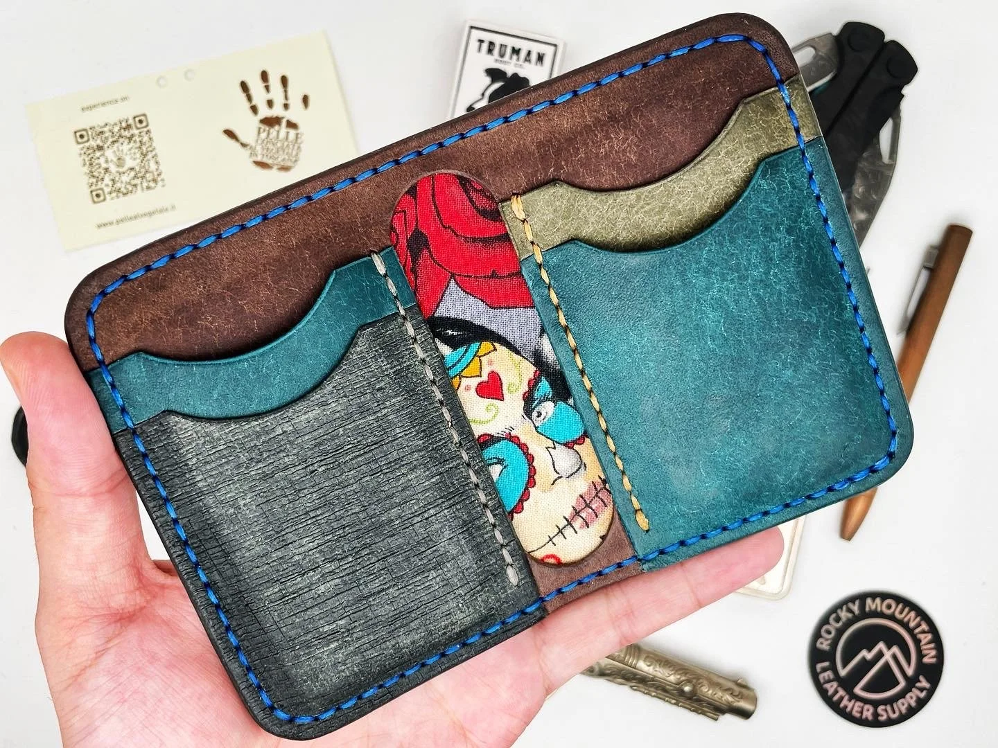 Colorful leather wallet with multiple compartments, featuring a vibrant skull with flowers design, held in hand, alongside various accessories and a Rocky Mountain Leather Supply sticker on a white surface.