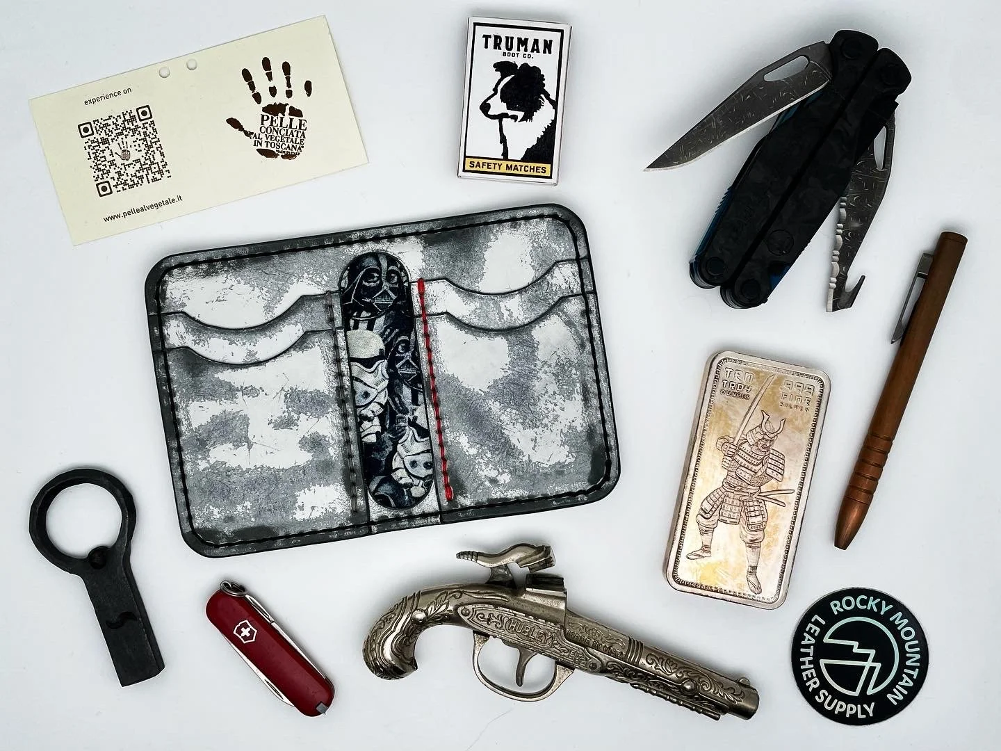 Arrangement of various tools and items on a white surface, including a Swiss Army knife, a brass pistol, a patch with a Rocky Mountain Leather Supply logo, a gold-colored card with a samurai illustration, a pocket pen, a multi-tool, a small echo of a