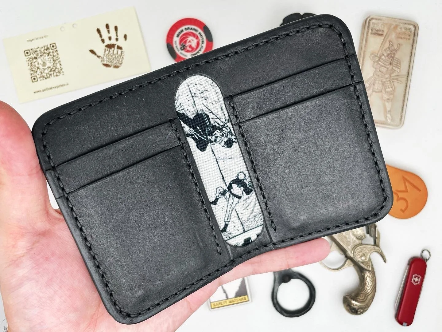Black leather wallet with a comic strip strip in the center, surrounded by various small objects including a white card with a QR code, a red and black poker chip, a silver gun-shaped keychain, a bottle opener, a small carved figure, a Swiss Army kni