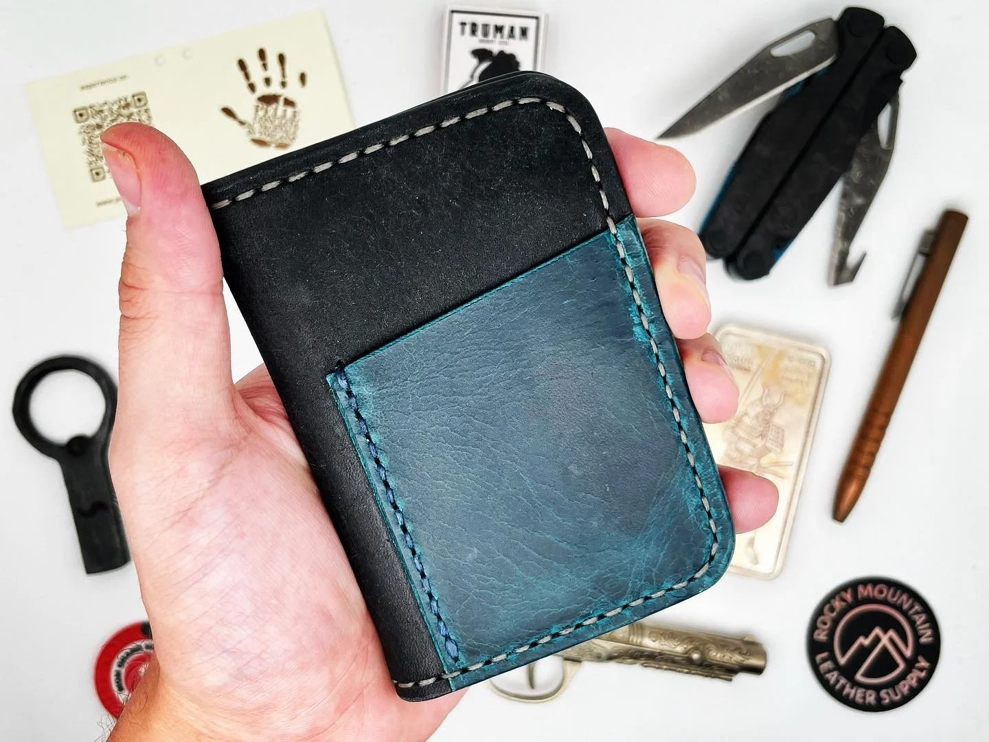 A hand holding a black and blue leather wallet with visible stitching, placed over a white surface with various tools, a sticker, a key, a pen, a card, and other miscellaneous items.