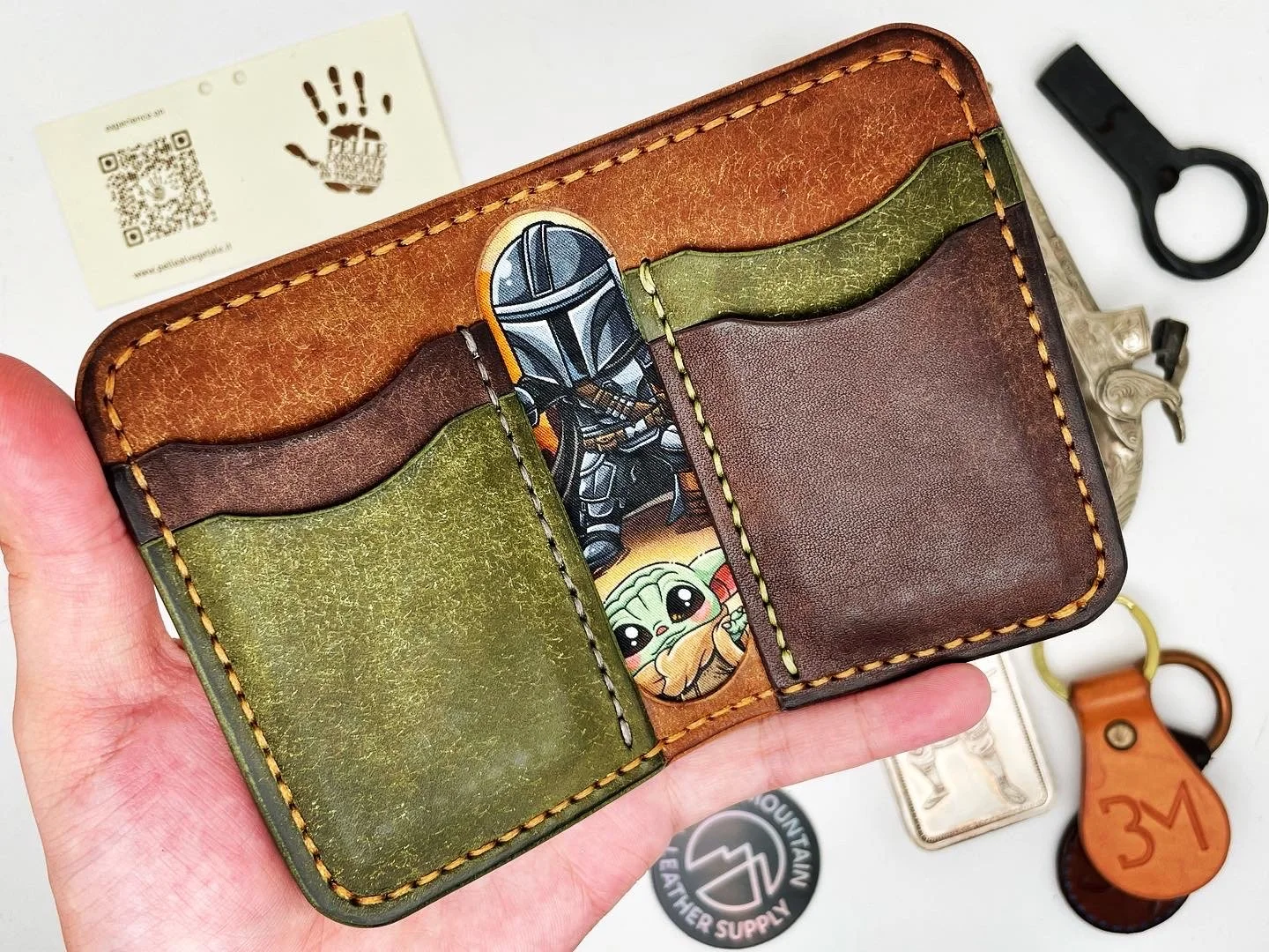 A multicolored leather wallet held in a person's hand, featuring a Star Wars stallone and Baby Yoda illustrations. Various items including a business card, keychains, and a metal emblem are laid out on a white surface in the background.