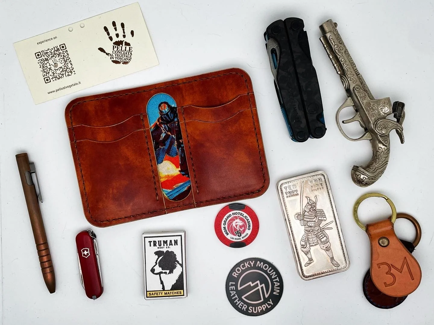 Collection of miscellaneous items including a brown leather wallet, a black multi-tool, a decorative silver toy gun, a silver bar with a samurai illustration, a round badge, a keychain with a leather strap and 3M logo, a red Swiss Army knife, a gold 