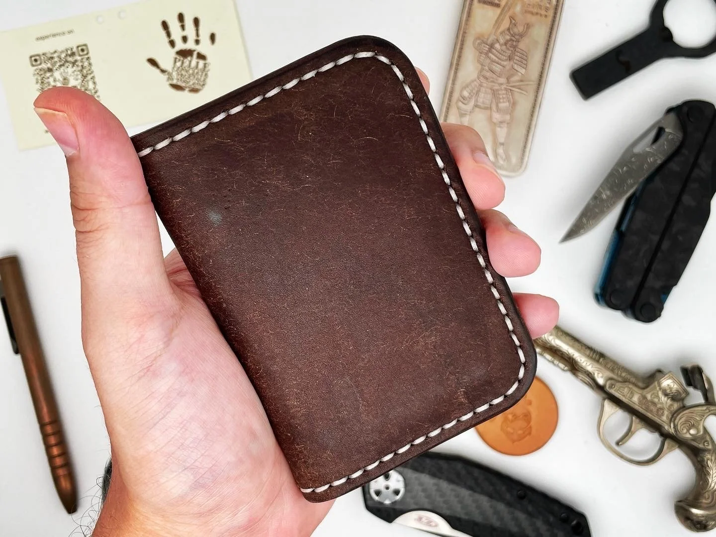 A person's hand holding a brown leather wallet. On the table beneath are various objects including a poker chip, a gold-colored handgun with ornate details, a black and silver knife, a Swiss army knife, a small flashlight, a lighter, a tiny black fla