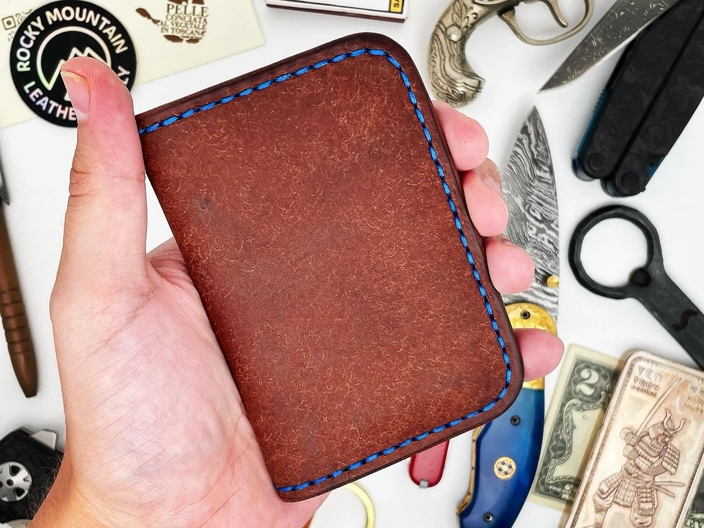 A hand holding a brown leather wallet with blue stitching, surrounded by various knives, a flashlight, a pair of scissors, a wallet, and dollar bills on a white surface.