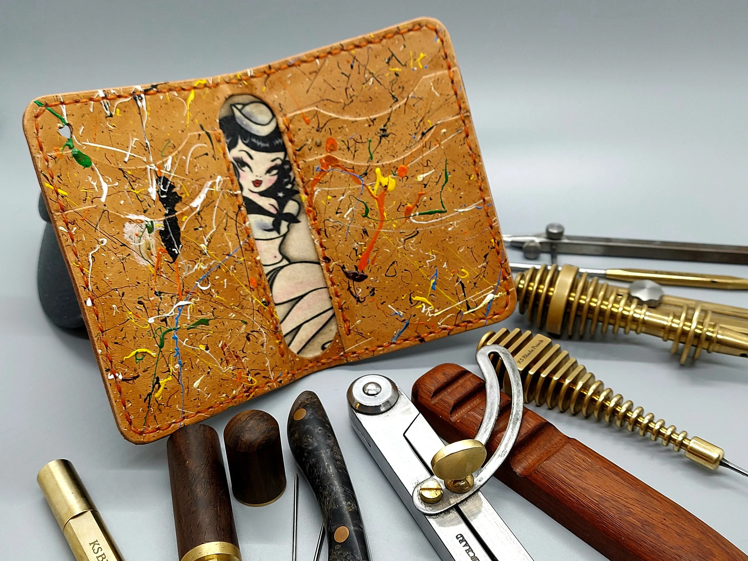 A decorated leather wallet with a pin-up style cartoon woman on the inside, surrounded by barber tools and accessories including straight razors, scissors, and blades on a light surface.