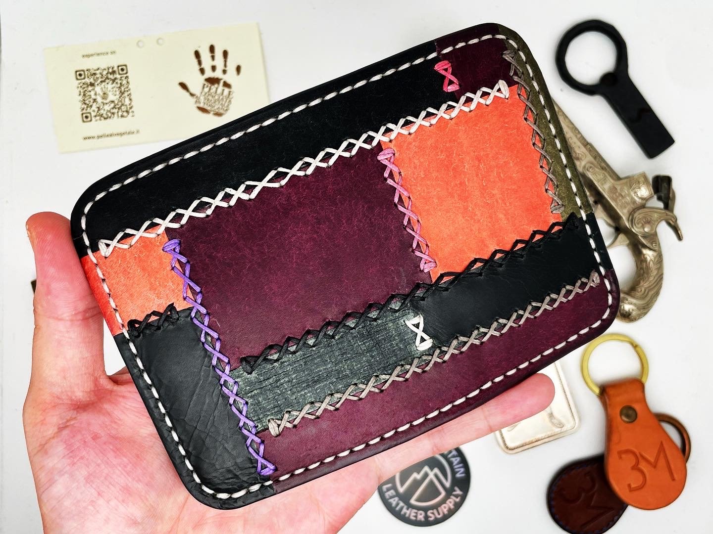 A hand holding a colorful leather wallet with patchwork design and decorative stitching. The wallet is black, purple, and orange with zigzag stitching. Surrounding items include keys and a business card with a handprint logo.
