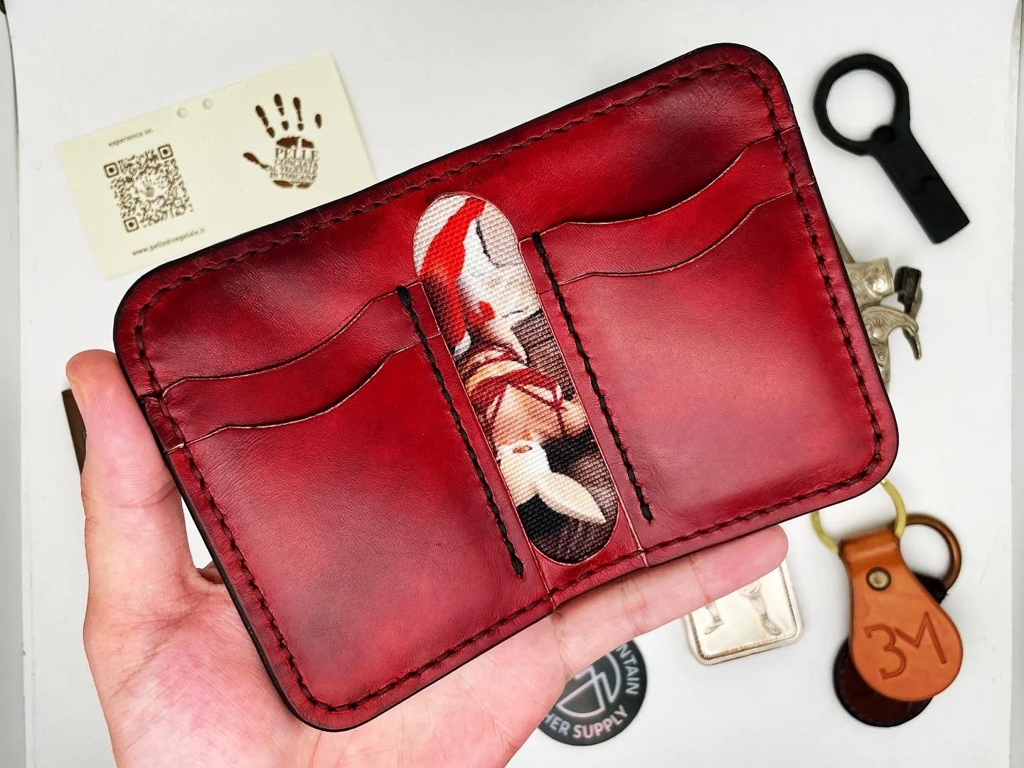 Red leather wallet with multiple card slots, featuring a vertical fabric strip with a woman’s face on it, held in a person's hand. Various items including a black keyring, a bottle opener, a 3M keychain, and a business card are on the surface.