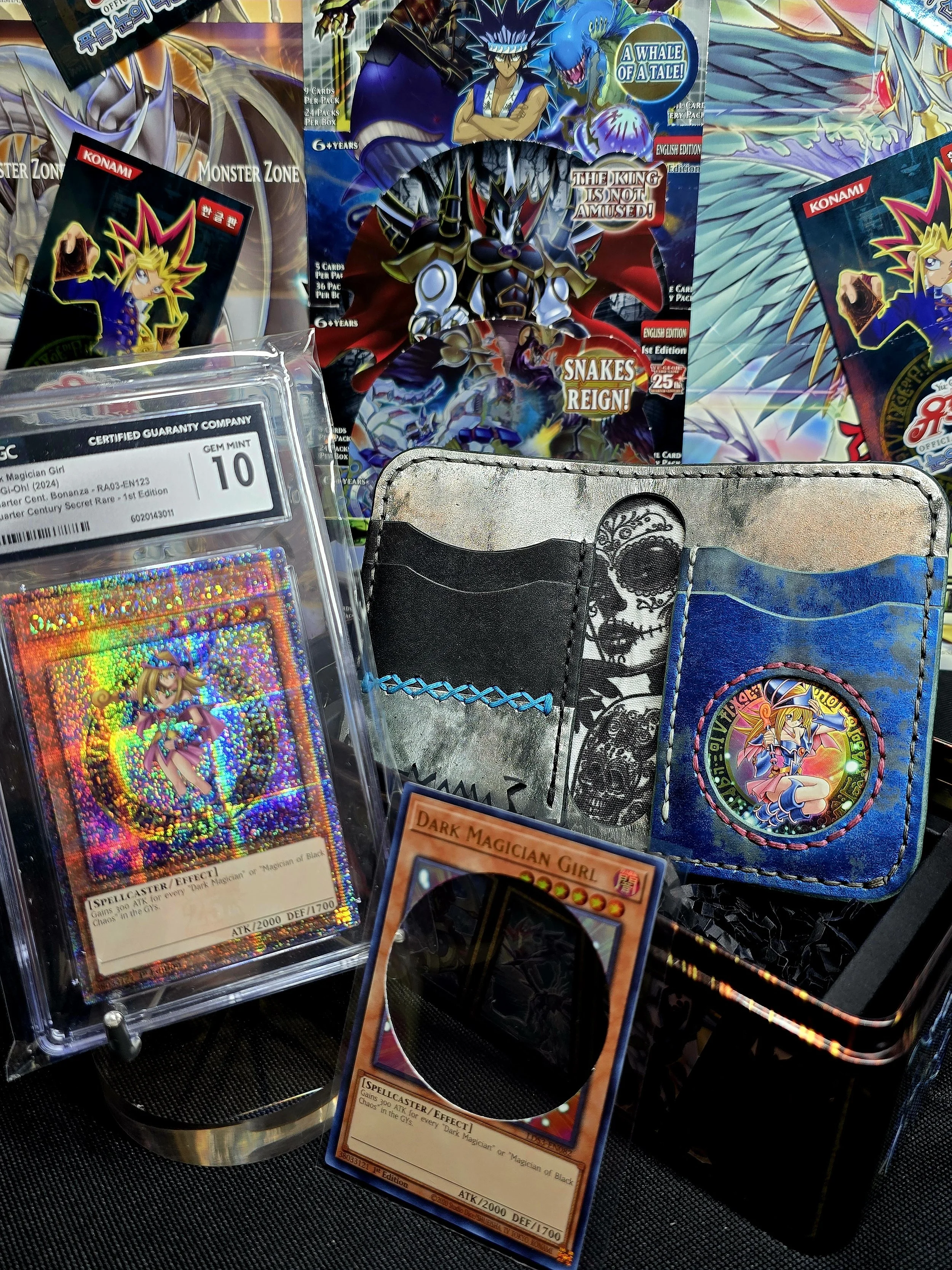 Collection of Yu-Gi-Oh! trading cards, including a holographic Dark Magician Girl card, with Yu-Gi-Oh! themed merchandise, booster packs, and a wallet with skull art in the background, displayed on a black surface.