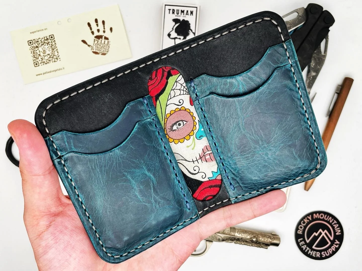 A hand holding a leather wallet with a colorful, artistic tattoo design on the inside. The wallet is blue with white stitching, and there are card slots visible. Various objects are surrounding the wallet, including business cards, a badge, a pen, an