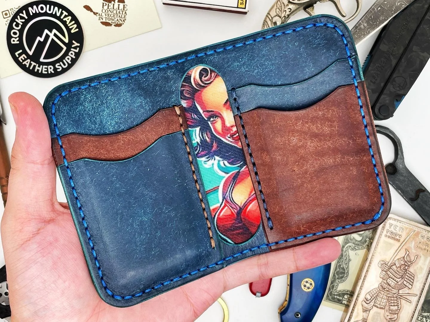 A hand holding a leather wallet with card slots, featuring a colorful cartoon-style image of a woman. The wallet has blue and brown leather sections with blue stitching. Surrounding the wallet are various knives, a book or box, and U.S. dollar bills 