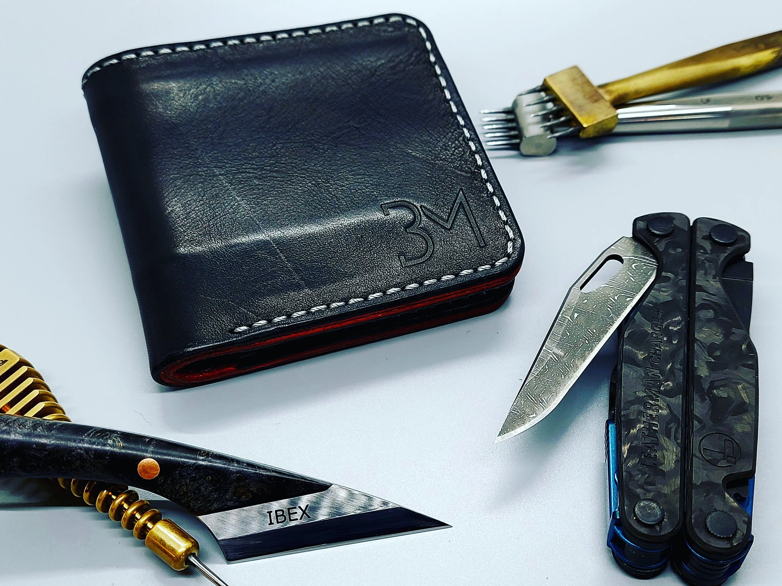 A black leather wallet with white stitching, a gold-handled hair clip, an IBEX straight razor, and a multitool with a bottle opener on a white surface.