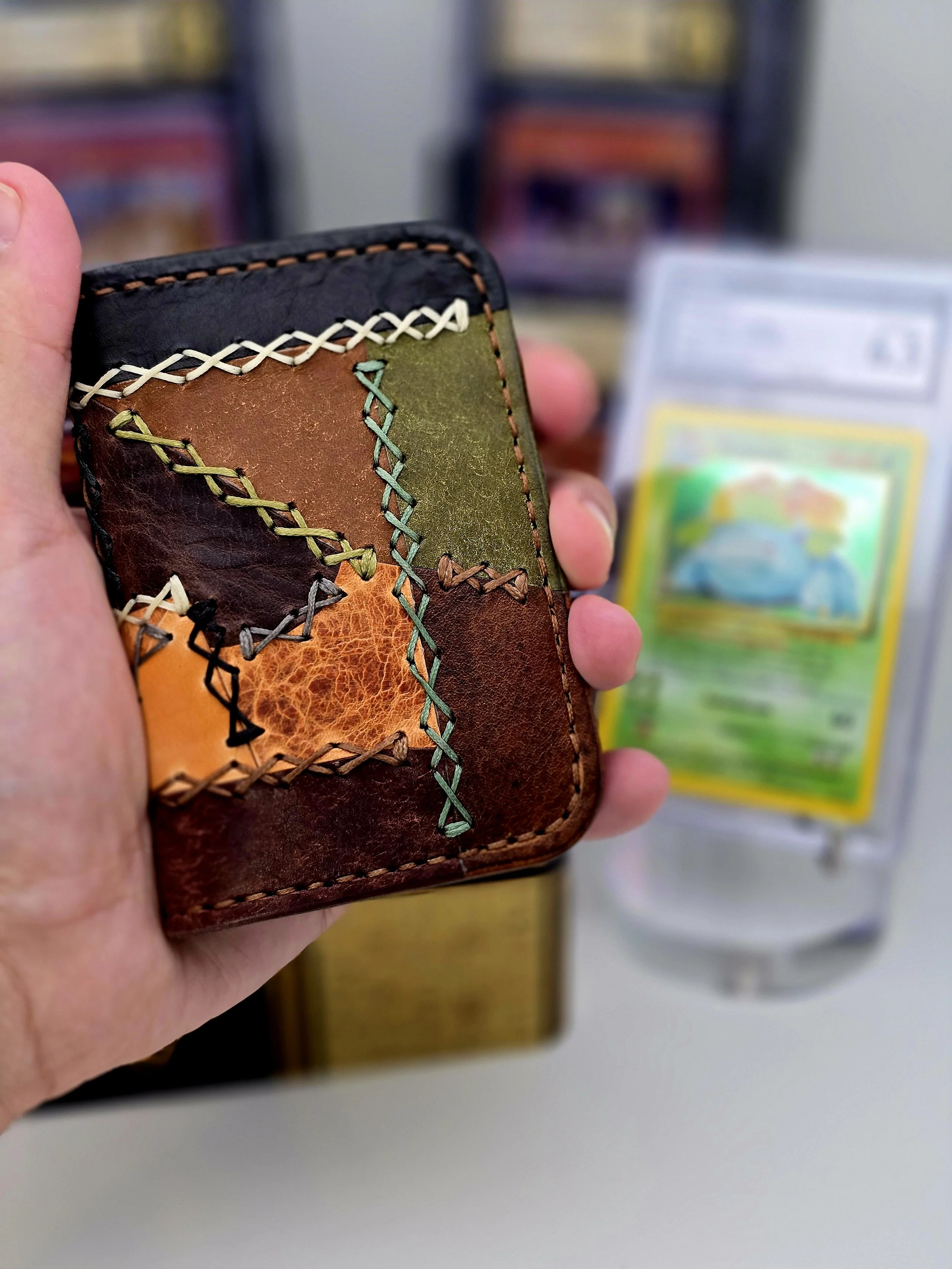 Colorful patchwork leather wallet with visible stitching, held in a hand with a blurred background that includes a Pokémon card in a plastic case.