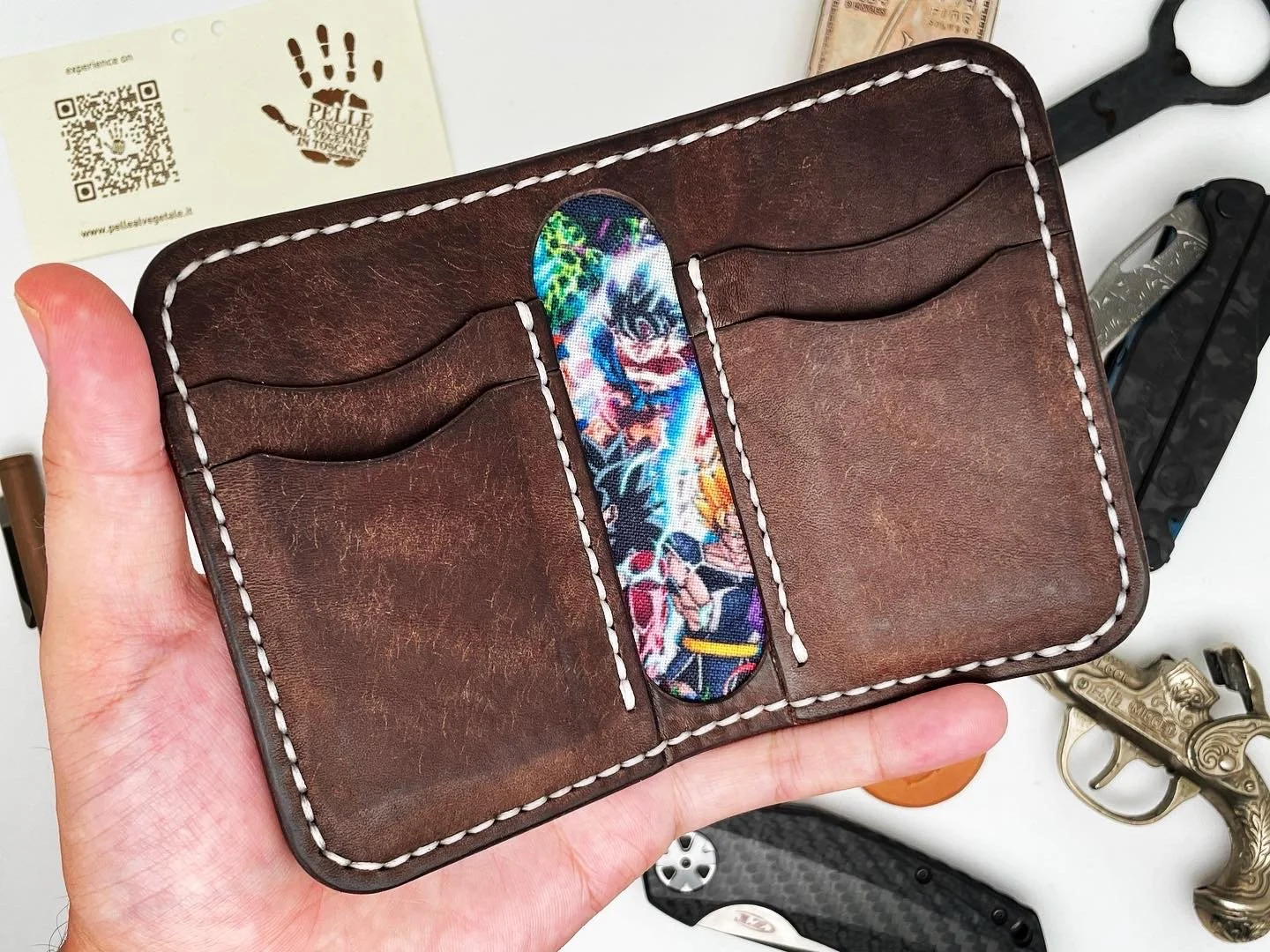Brown leather wallet with white stitching, three card slots, and a colorful fabric card in the middle slot. Hand holding the wallet, objects on a white surface nearby including a business card, a folding knife, and a small toy gun.