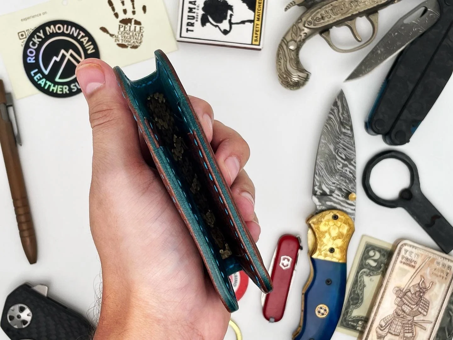 A person holding an open, empty leather wallet over a cluttered white surface with various knives, a Swiss Army knife, cash, a utility knife, and stickers scattered around.