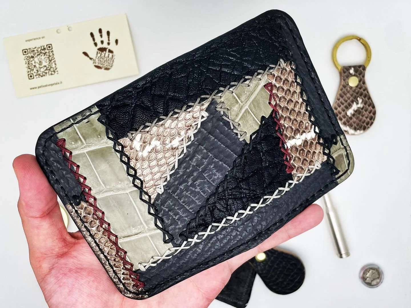 A hand holding a rectangular-shaped cardholder made of snakeskin-patterned material with red and white stitching, with a black leather border and grid pattern on the inside.