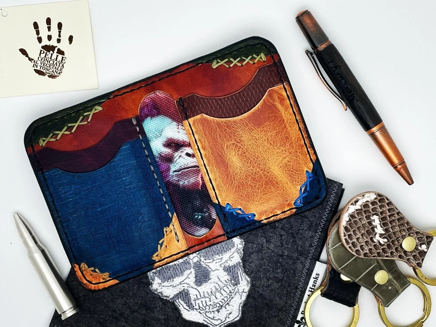 A colorful wallet with a portrait of a smiling woman, surrounded by pens, scissors, a skull-printed cloth, and a small white note with a black handprint and text in Italian.