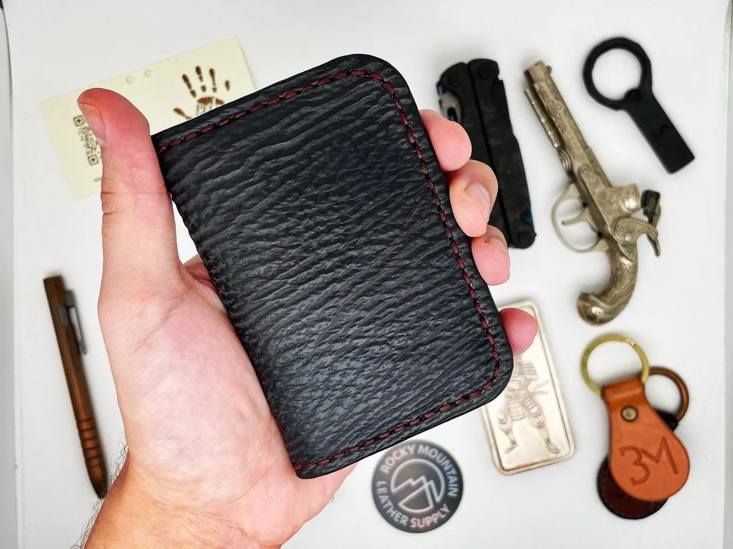 Hand holding a black textured wallet with red stitching, with various small items on a white surface in the background, including a miniature pistol, keychains, and a button.