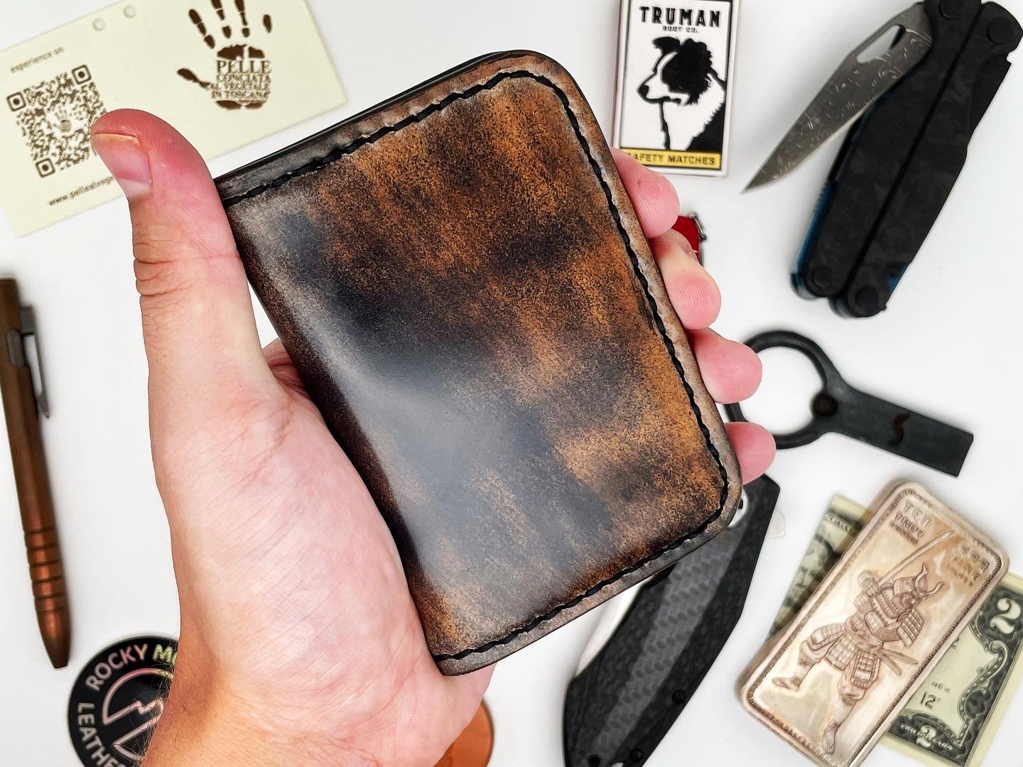 Hand holding a worn brown leather wallet with visible stitching, surrounded by various items including a multi-tool, U.S. bills, a pocket knife, a keychain, a matchbox, and a card with a QR code.