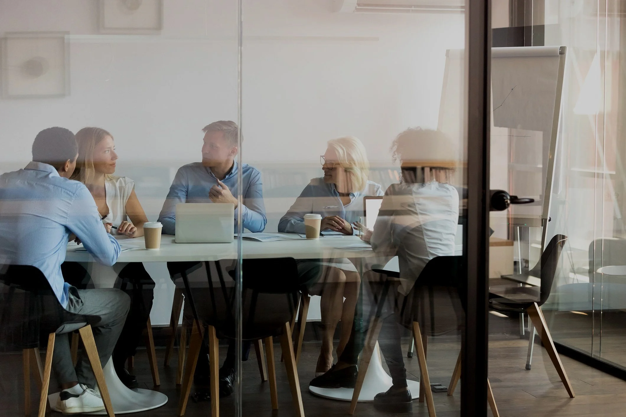 A group of five people having a meeting in a conference room, seen through glass with a flip chart in the background, and coffee cups on the table.