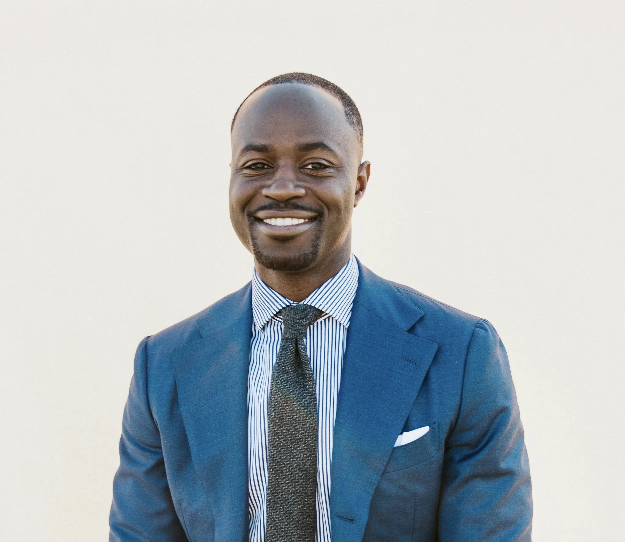 Smiling man in a blue suit, dress shirt with blue and white stripes, and a textured tie, standing against a light background.